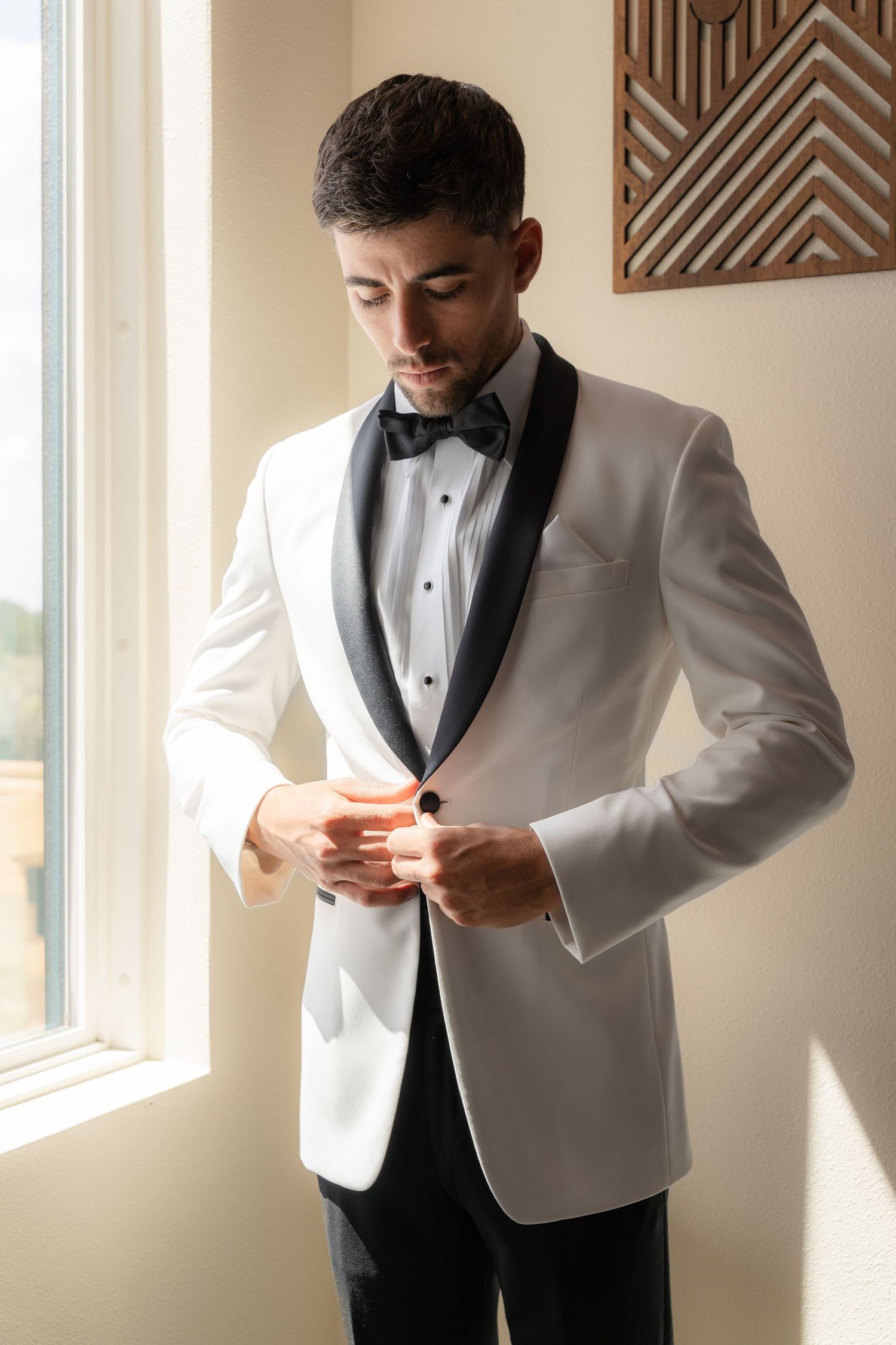A groom in a white tuxedo jacket with black lapels adjusts his cufflinks by a sunlit window.