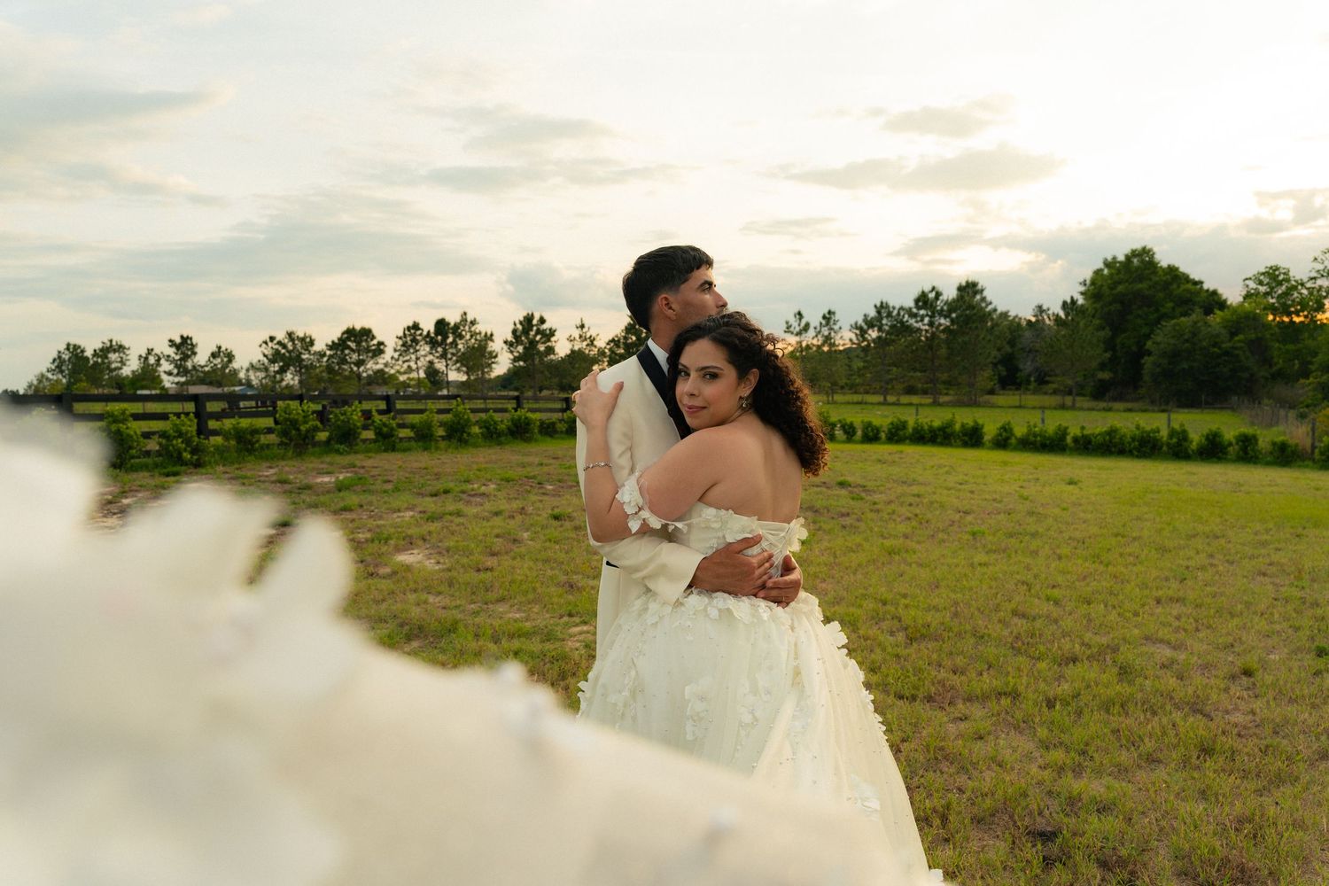 Bride and groom embrace in a green field at sunset with veil flowing in the foreground.