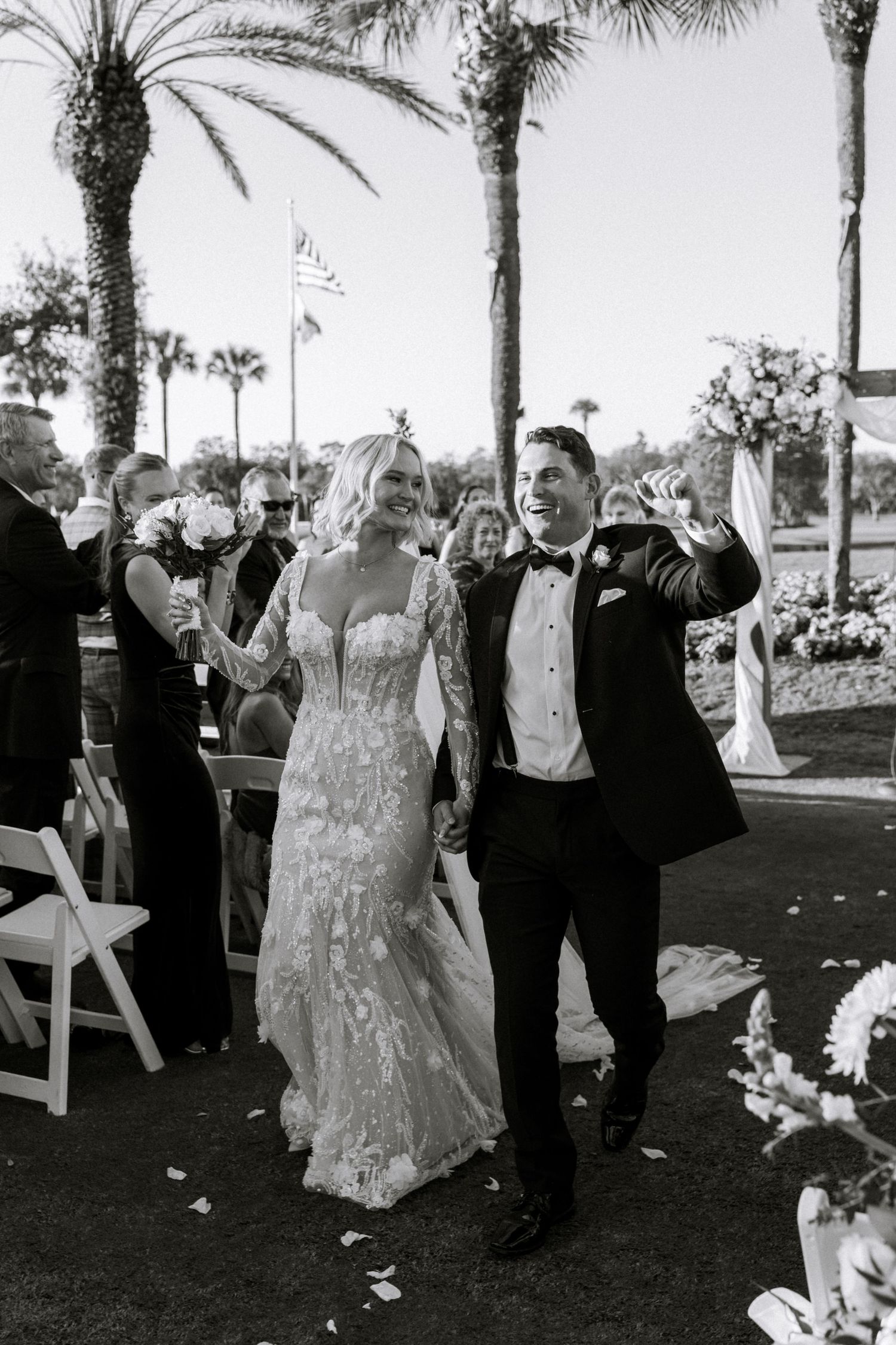 Joyful newlyweds walk down the aisle after wedding ceremony outdoors near palm trees in black and white photo.
