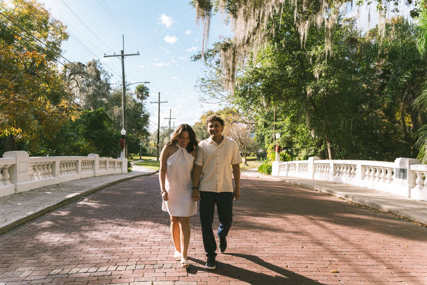 A couple walks together on a brick bridge lined with Spanish moss and palm trees on a sunny afternoon.