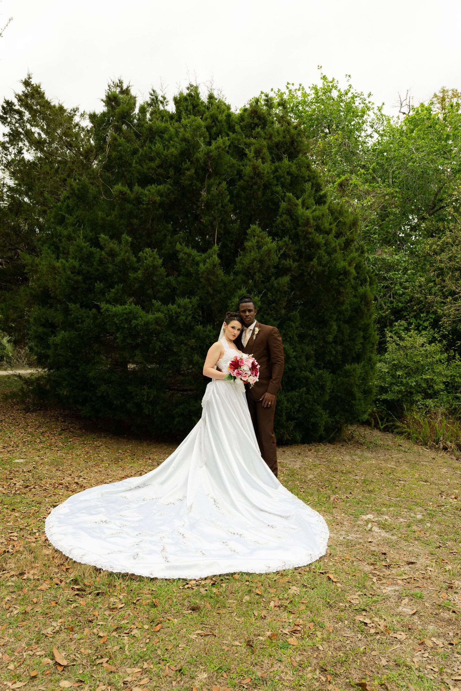 Bride in white gown with long train stands with groom in brown suit before evergreen trees.