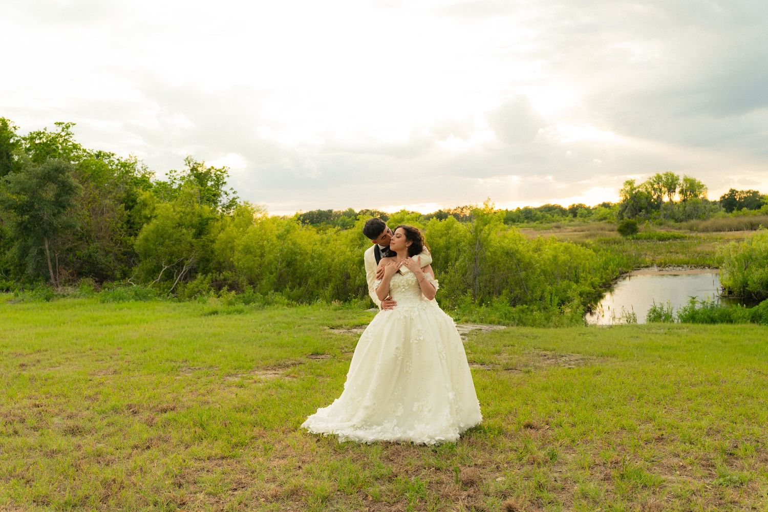 Groom kisses bride in a lace ball gown beside a serene pond surrounded by lush greenery.