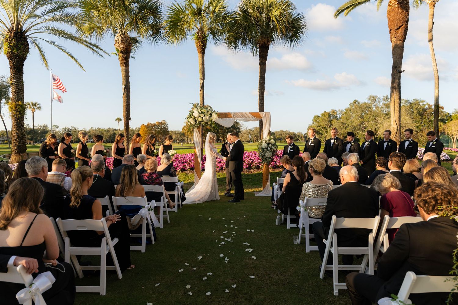 Outdoor wedding ceremony on a golf course with guests seated in white chairs under palm trees.