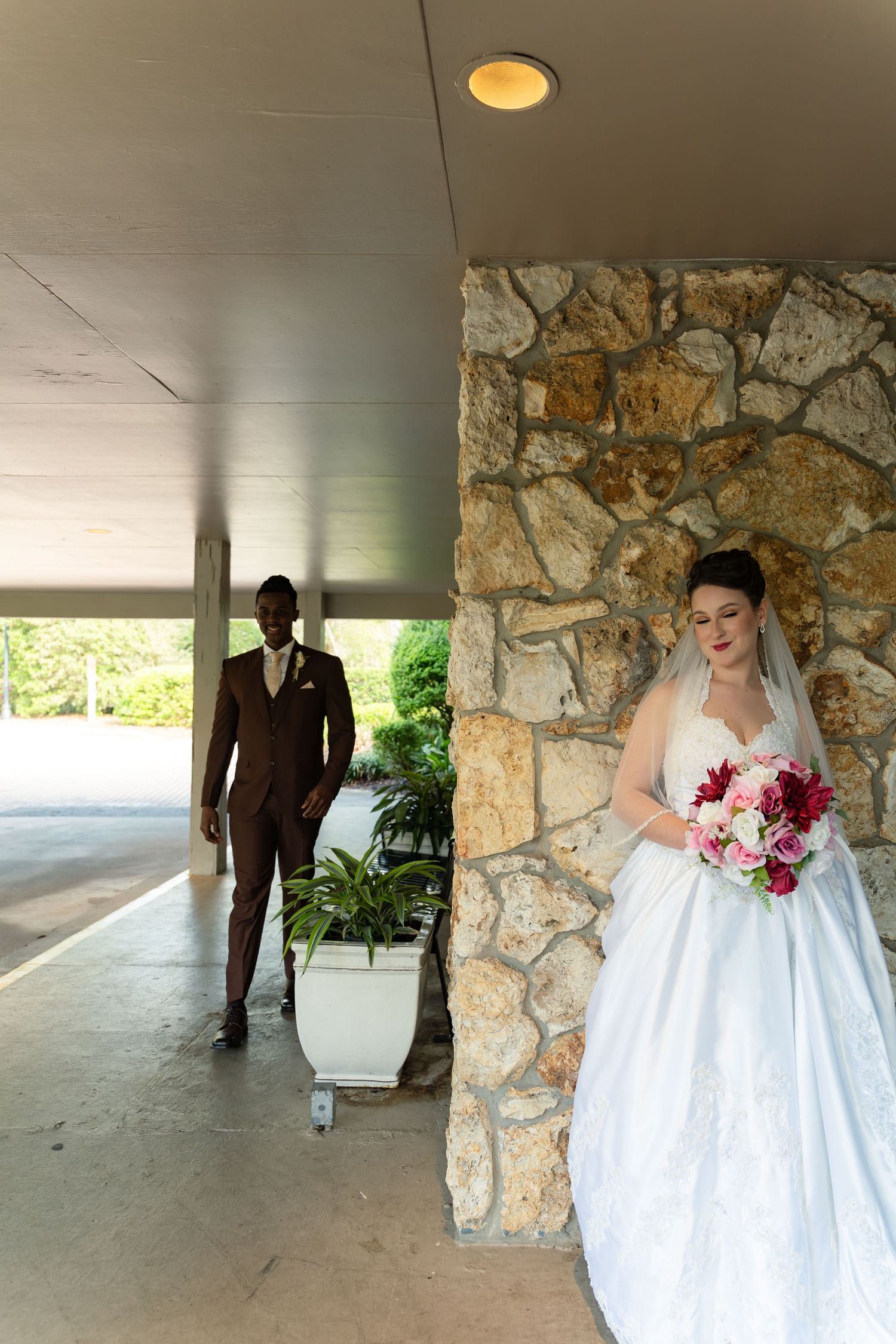 Bride holding bouquet stands by stone wall as groom approaches in brown suit during wedding first look.