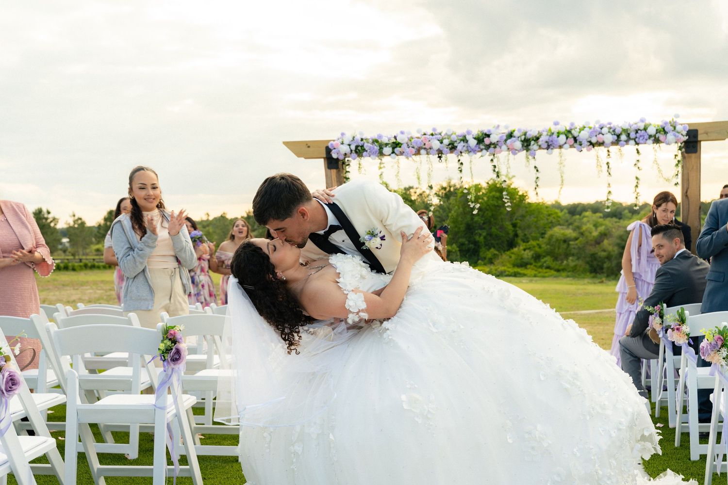 Groom dips bride in a romantic kiss at outdoor wedding ceremony with floral arch and guests watching.