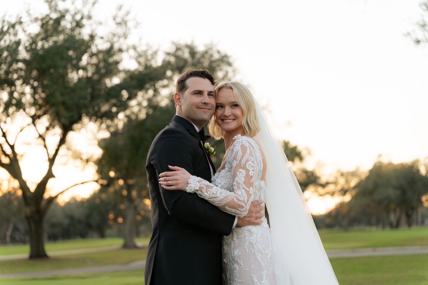 Bride and groom embrace outdoors at sunset, she in lace long-sleeve gown with veil, he in black tuxedo.