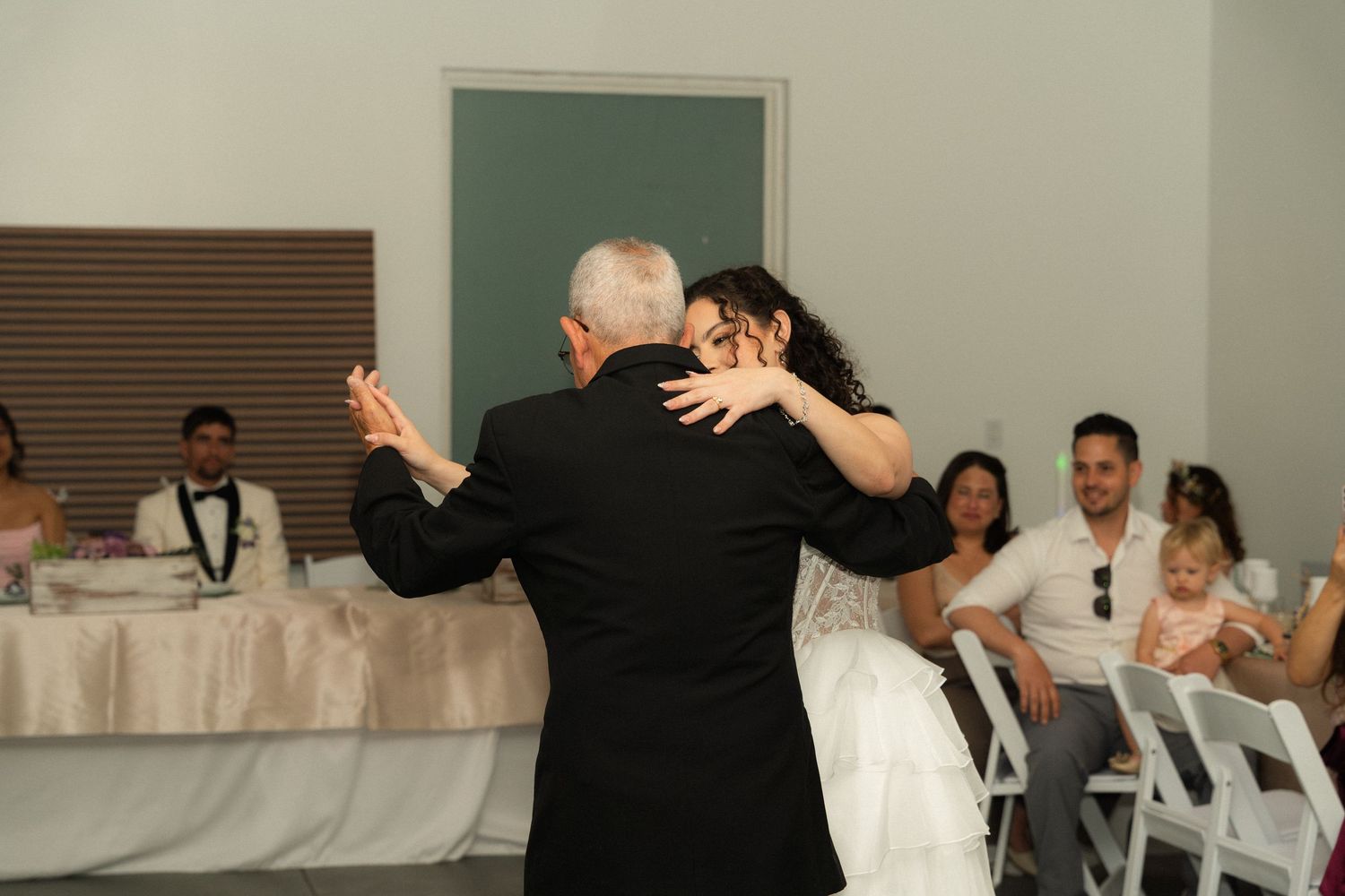 Bride shares an emotional father-daughter dance at wedding reception with seated guests watching nearby.