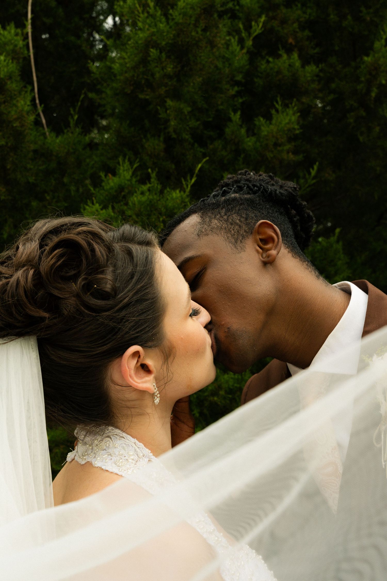 Interracial bride and groom share a romantic kiss outdoors, framed by a flowing white veil and dark green foliage.