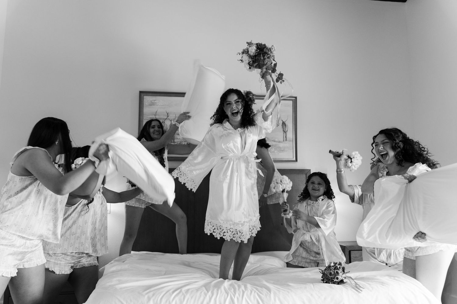 Bride and bridesmaids in white robes having a fun pillow fight on a bed during wedding morning preparations.