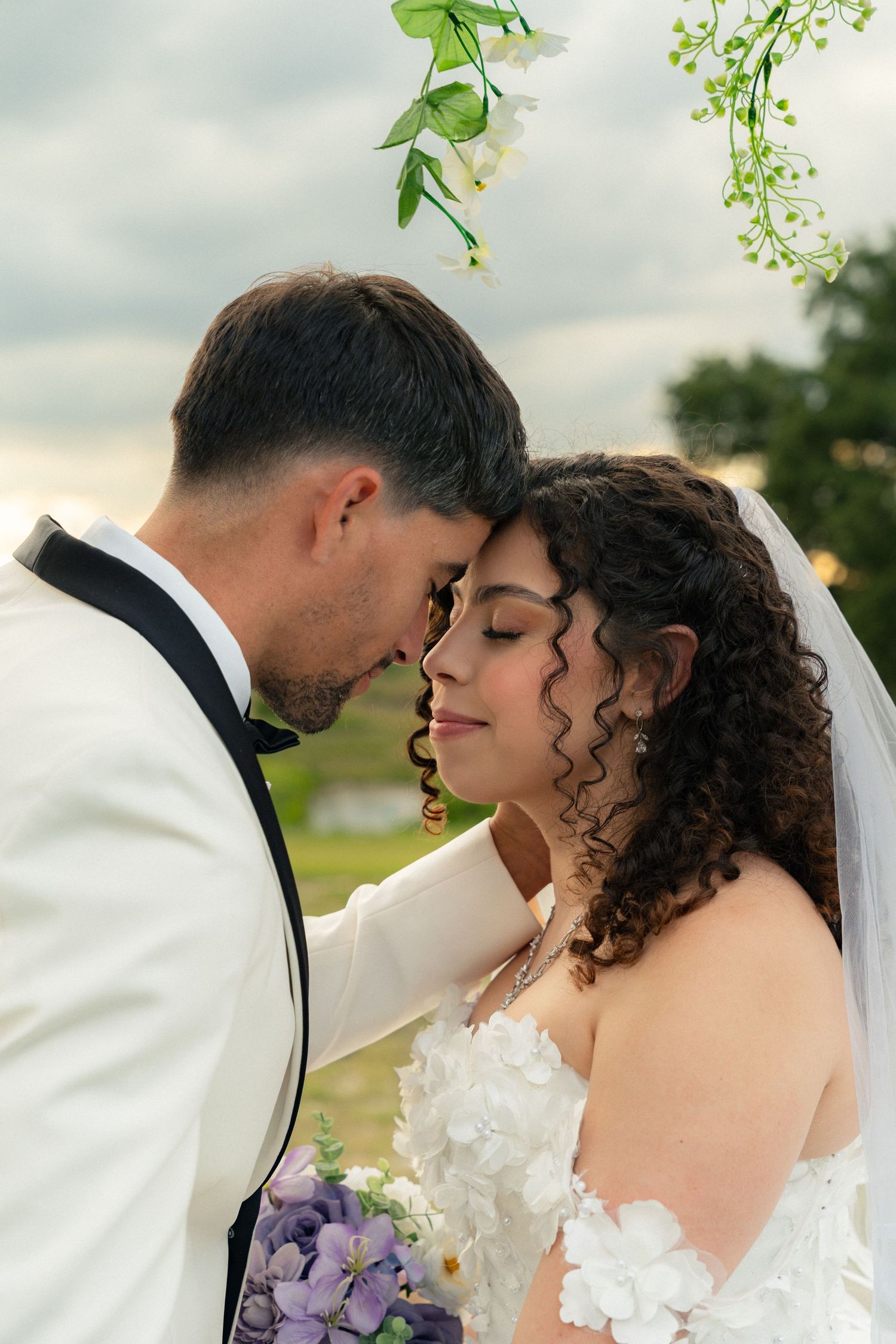 Bride and groom touching foreheads tenderly under floral arch at outdoor wedding ceremony.