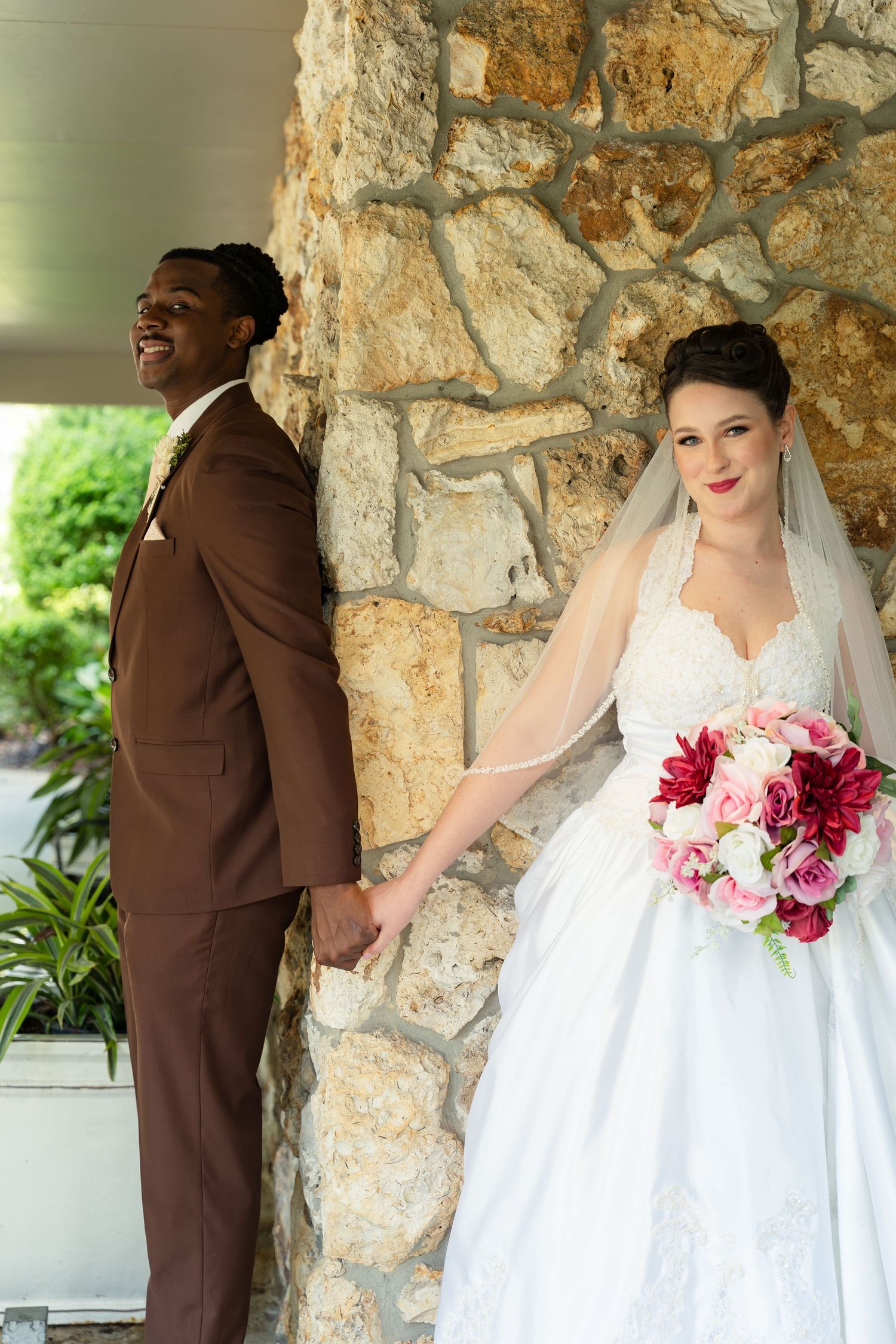Interracial couple poses back-to-back at stone pillar, groom in brown suit, bride in white gown holding pink bouquet.