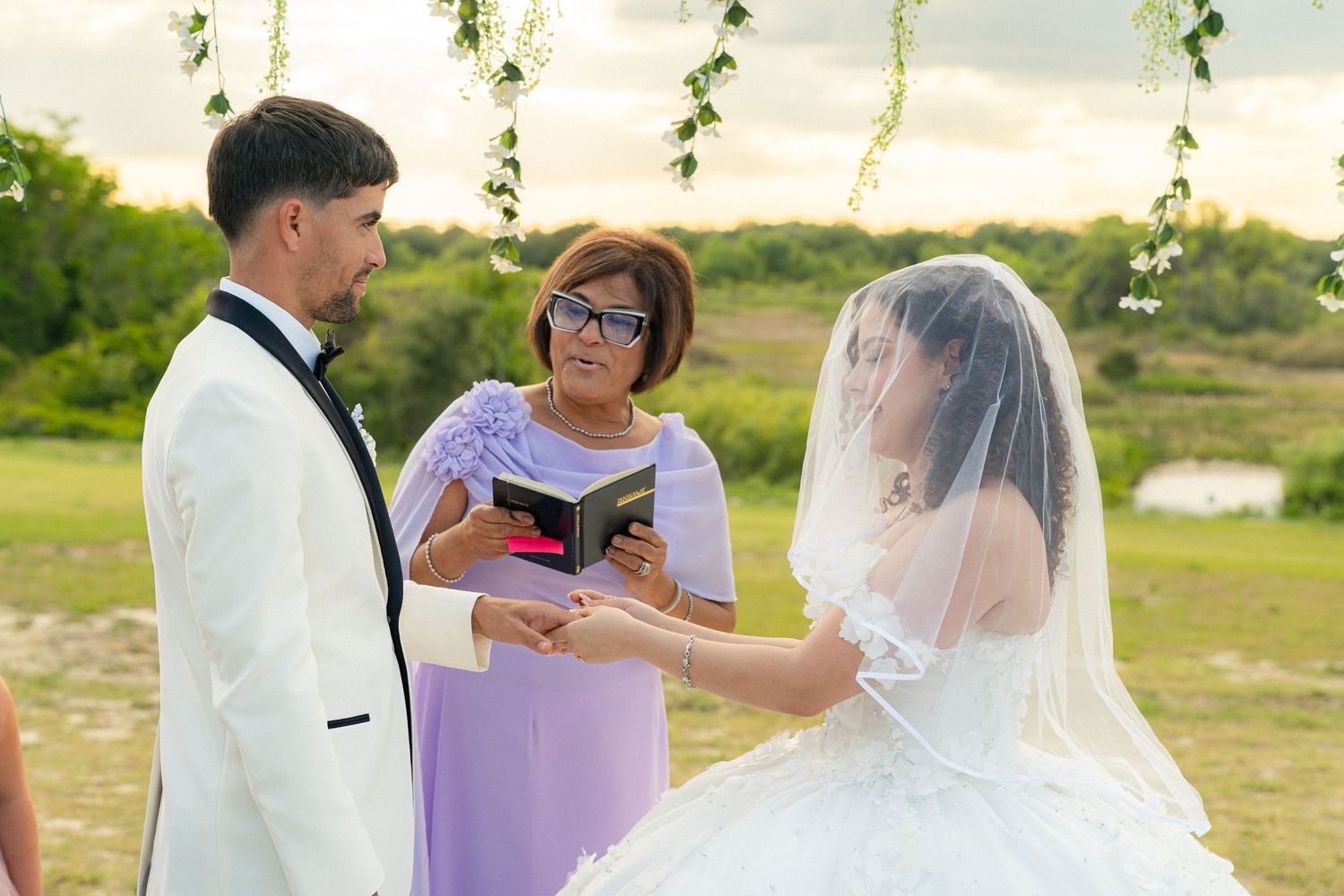 Outdoor wedding ceremony with bride in white gown and groom in white tuxedo exchanging vows with officiant.