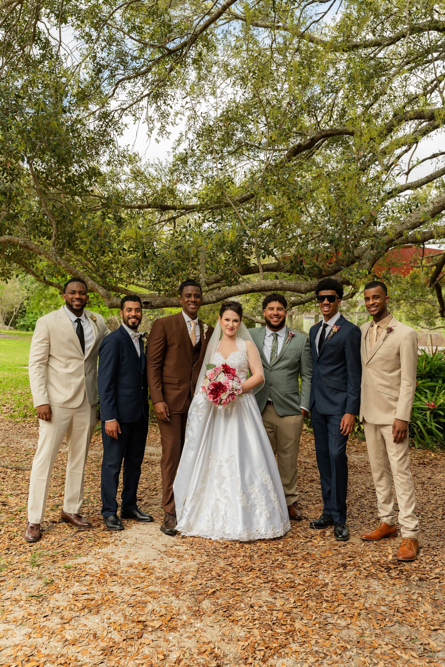 Bride in white gown poses with six groomsmen in suits under a large oak tree outdoors.