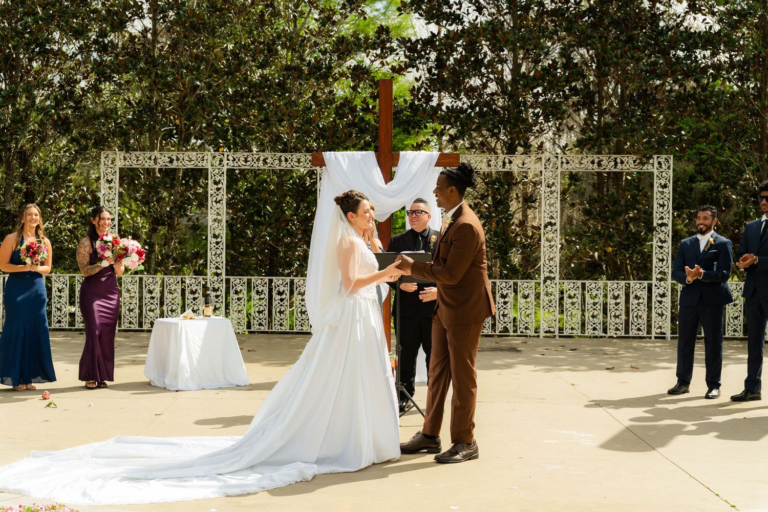 Bride and groom exchange vows at outdoor wedding ceremony with white lattice arch and wedding party standing behind them.