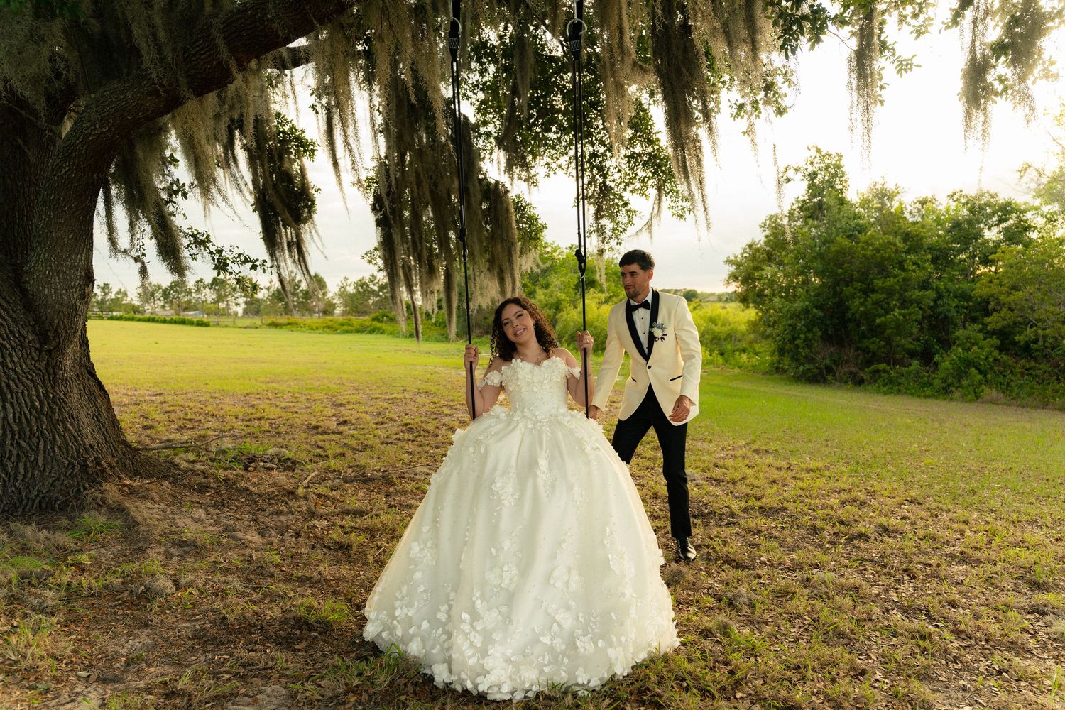 Bride in floral ball gown and groom in gold tuxedo posing under Spanish moss-draped oak tree.