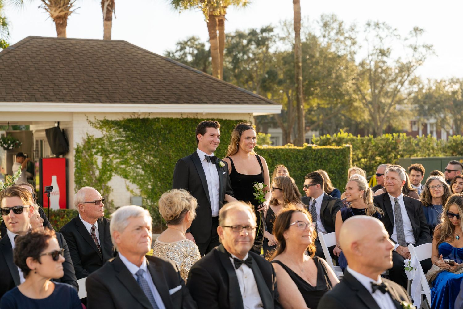 Elegant outdoor wedding ceremony with guests in formal attire seated on white chairs, surrounded by lush greenery and palm trees.