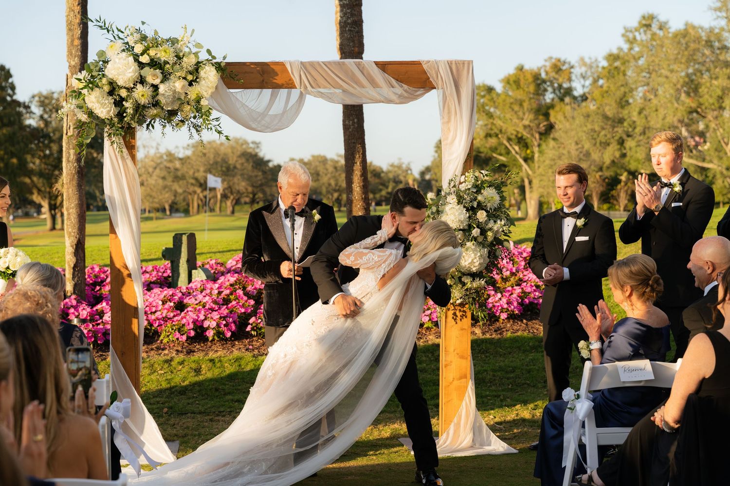 Bride and groom share first kiss under floral arch at outdoor wedding ceremony on lush golf course.
