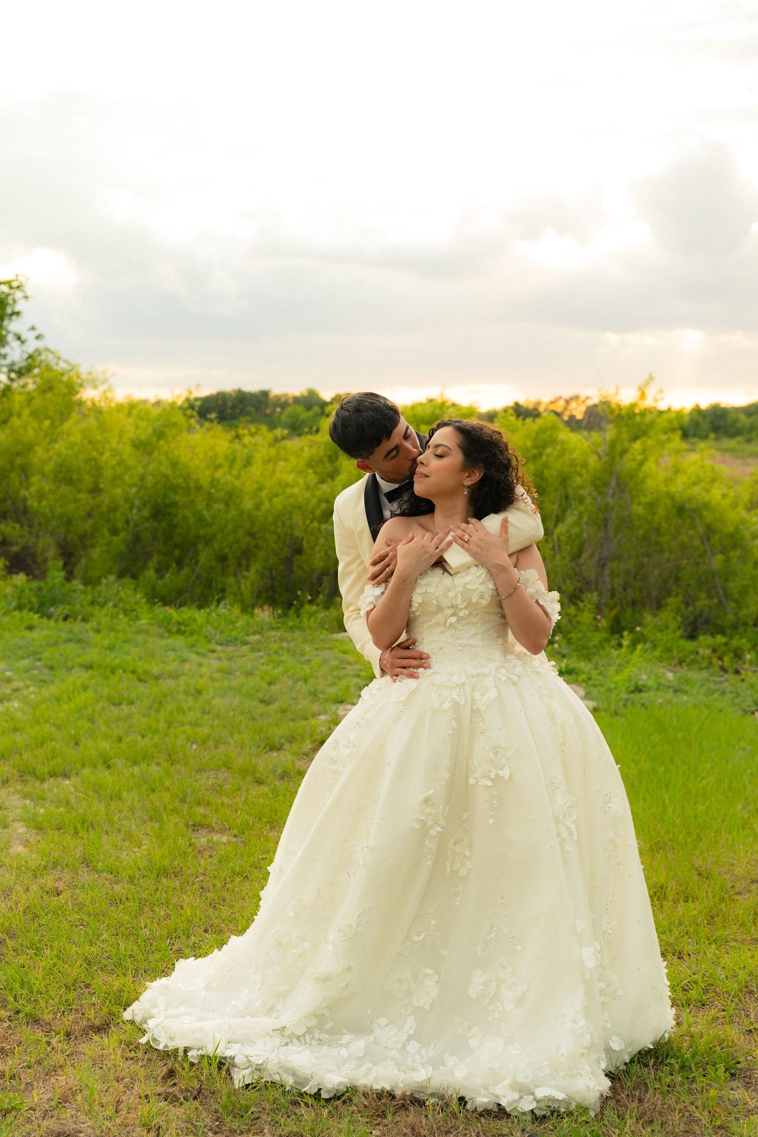 Groom in cream tuxedo embracing bride in floral ball gown during golden hour outdoor wedding.