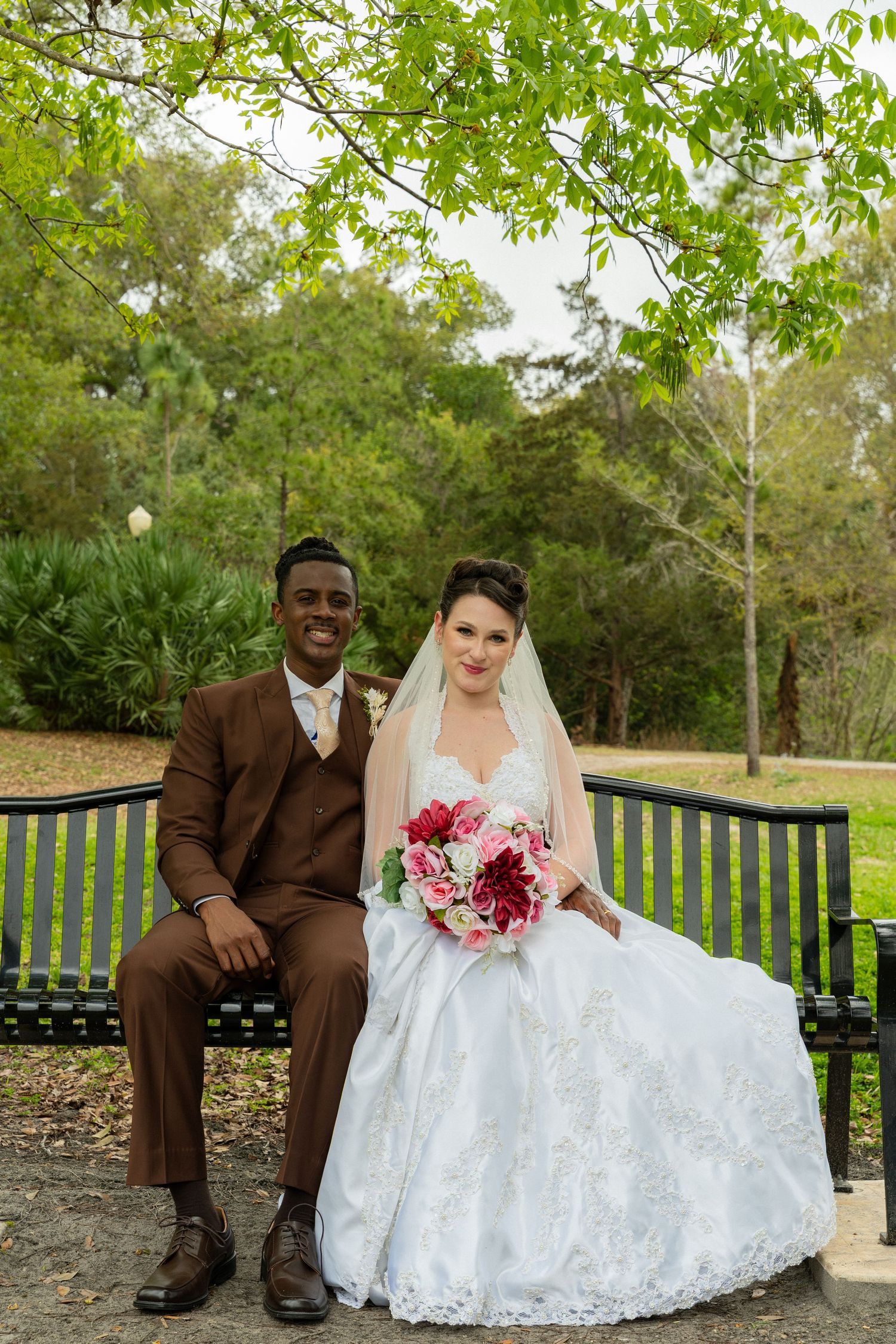 Smiling interracial newlyweds sit on a park bench surrounded by lush greenery, bride holding a pink and red bouquet.