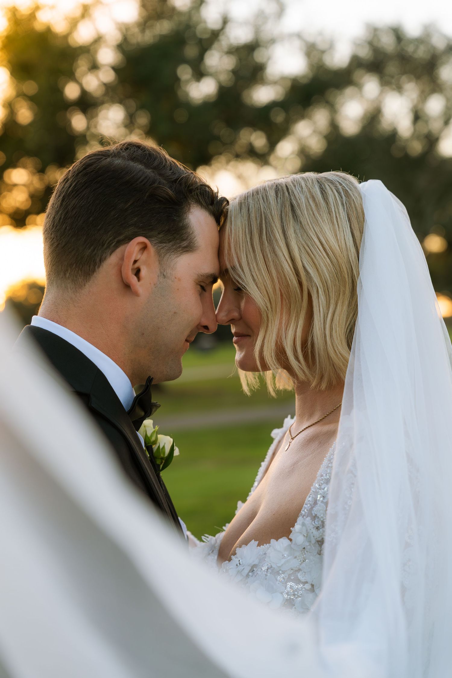 Bride and groom touch foreheads in a tender wedding moment outdoors at sunset, her veil flowing and a boutonniere on his tuxedo lapel.
