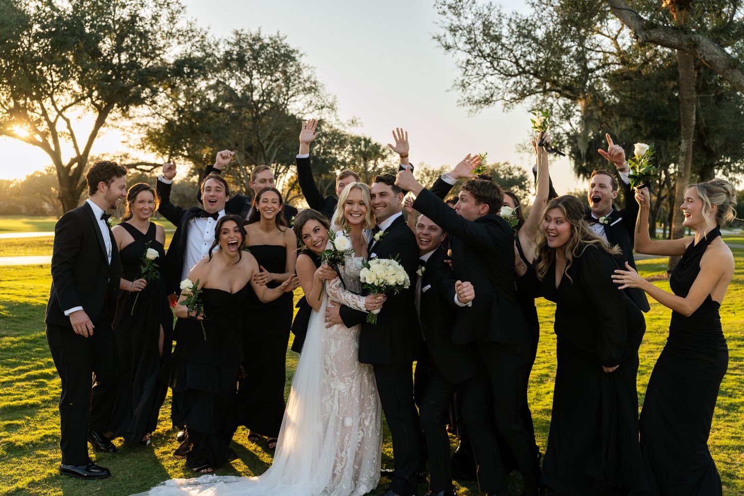 Bride in a lace wedding gown celebrates with cheering bridesmaids and groomsmen in black attire on a sunlit golf course lawn.
