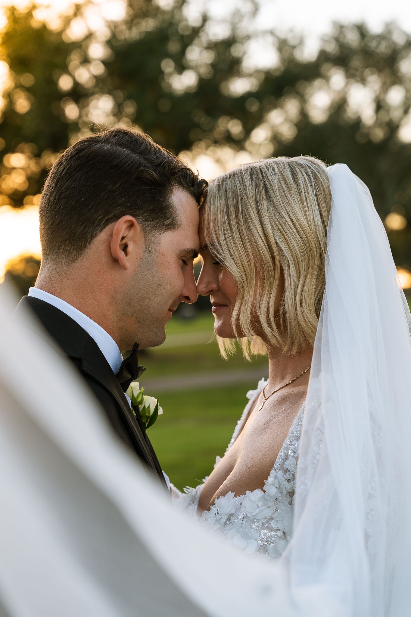 Intimate wedding portrait of newlyweds sharing a tender moment at sunset with bridal veil in foreground.