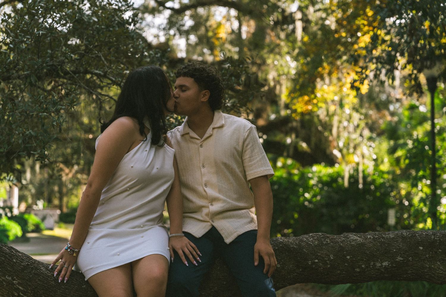 A couple in white outfits sits together on a tree branch in a sunlit outdoor setting during a romantic photoshoot.