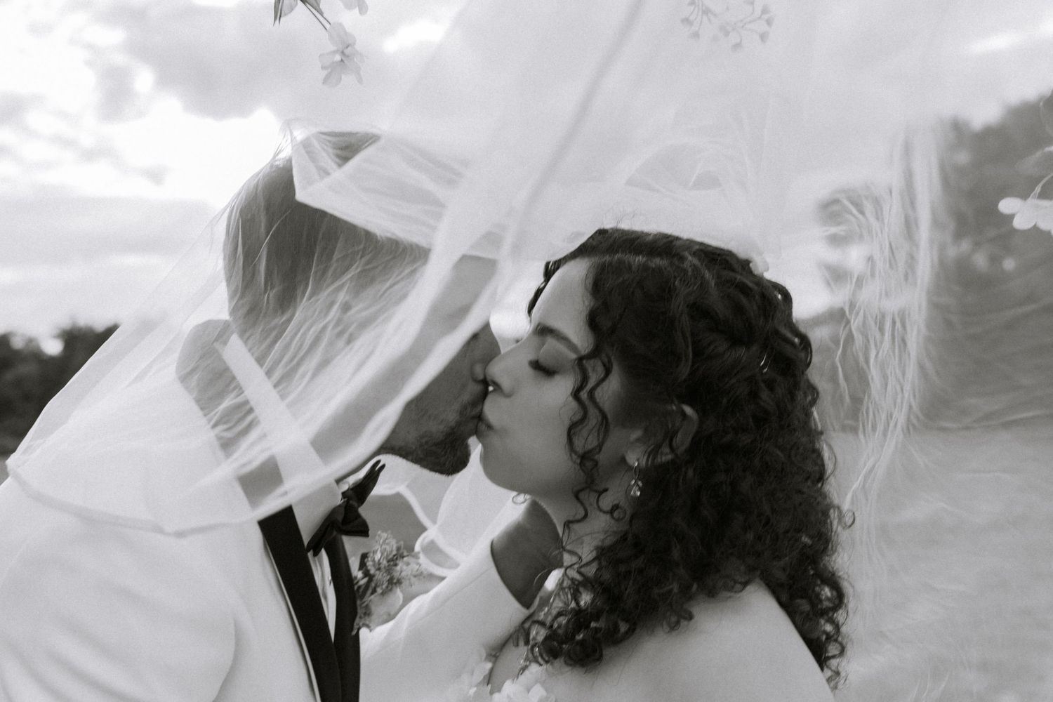 Black and white photo of bride and groom kissing under flowing wedding veil outdoors.