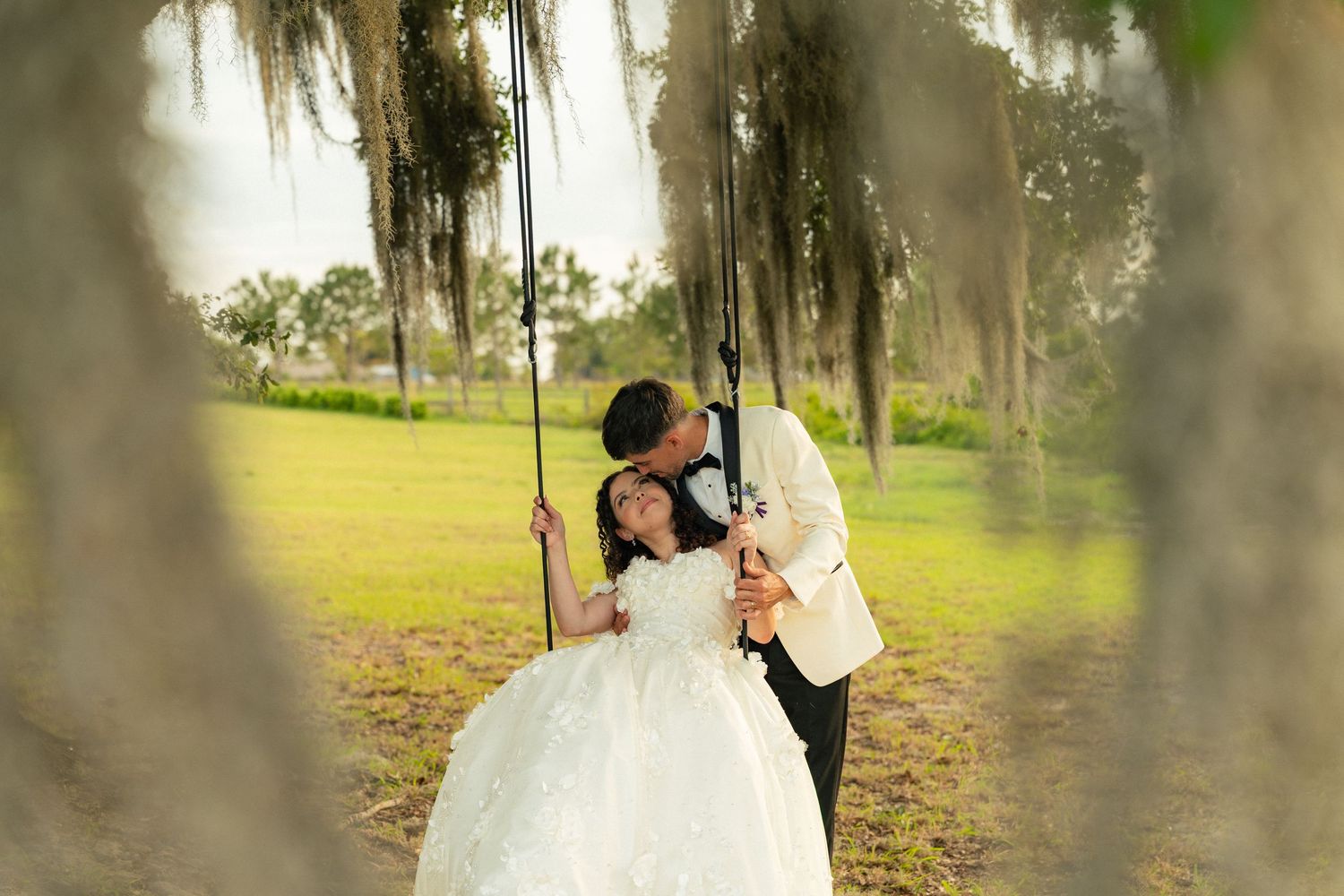 A groom kisses his bride sitting on a rope swing under Spanish moss-draped trees in a lush green outdoor setting.