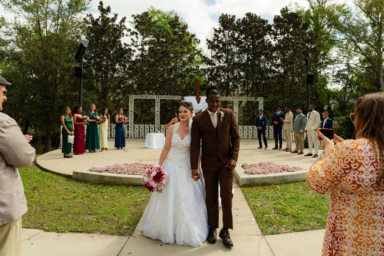 Bride and groom walk down aisle after outdoor wedding ceremony, wedding party standing behind them in colorful attire.