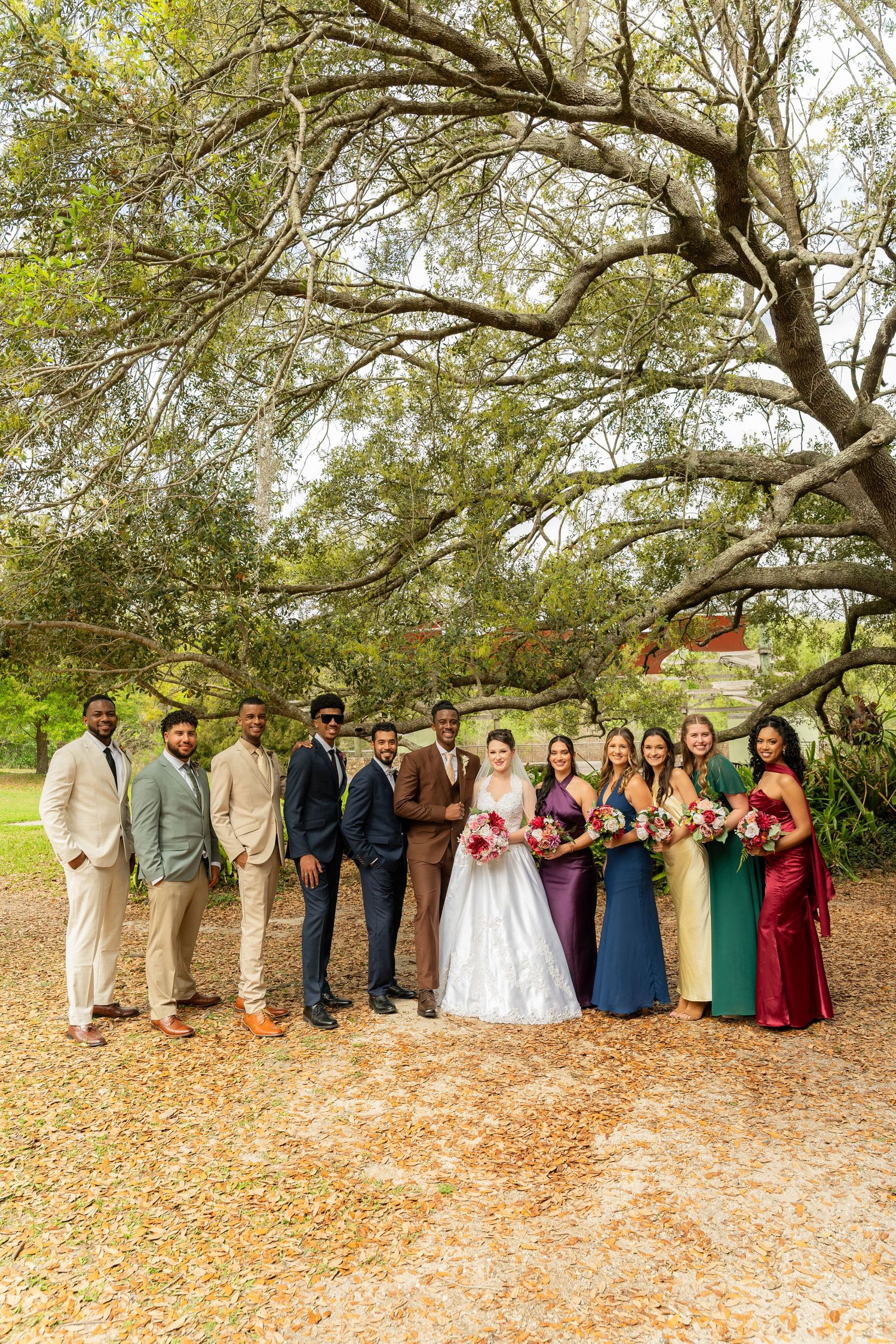 Wedding party poses under sprawling oak tree with bride in white gown and bridesmaids in jewel-tone dresses.