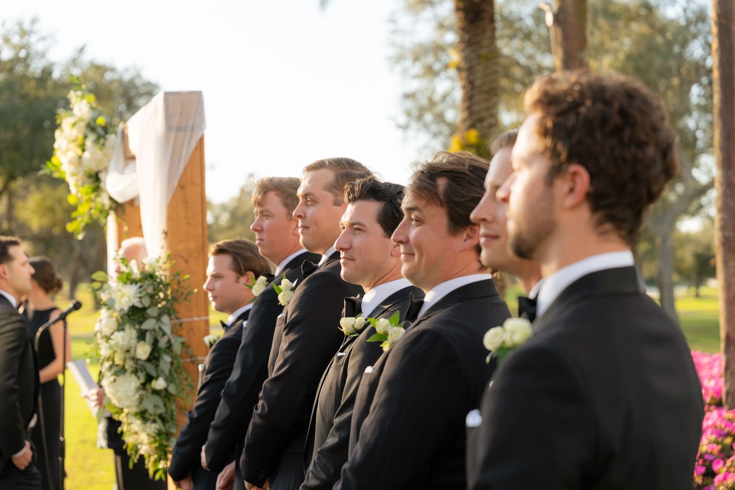 A row of groomsmen in black tuxedos smile during an outdoor wedding ceremony beside a floral arch.