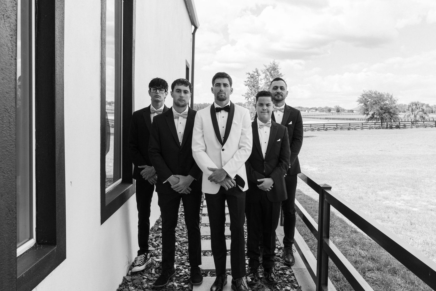 Black and white photo of groom in white tuxedo with four groomsmen in black suits posing outdoors.