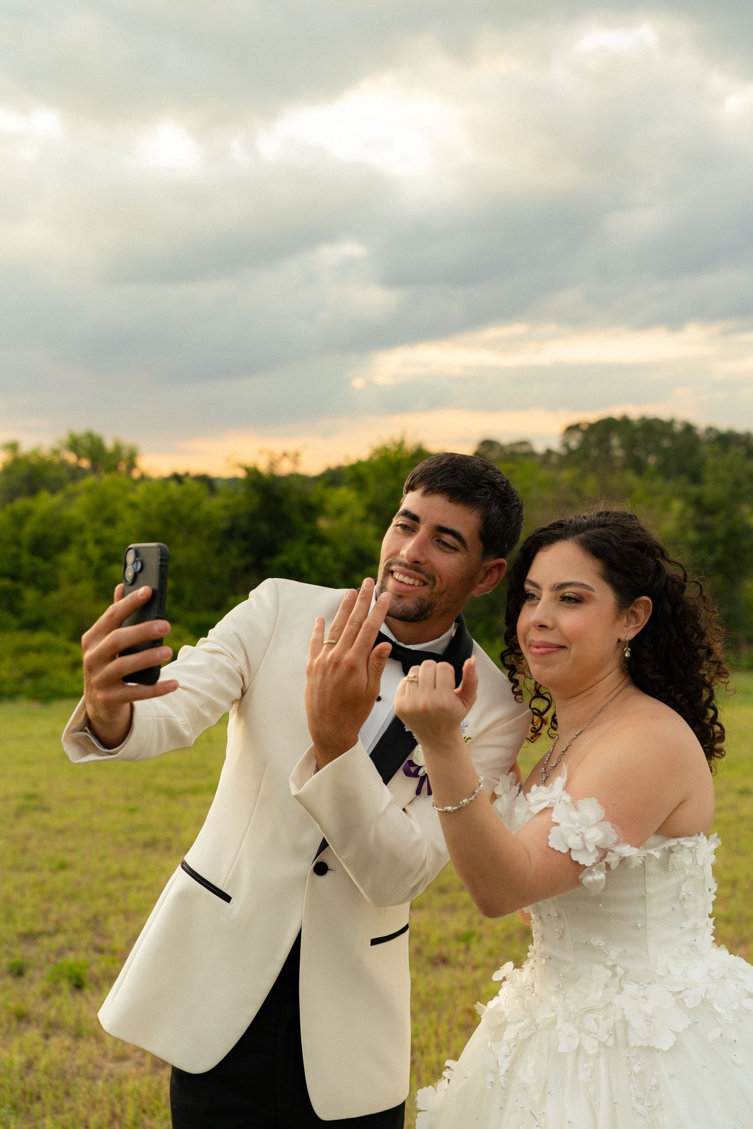 Smiling newlyweds taking a selfie outdoors at sunset, showing off their wedding rings in a field.