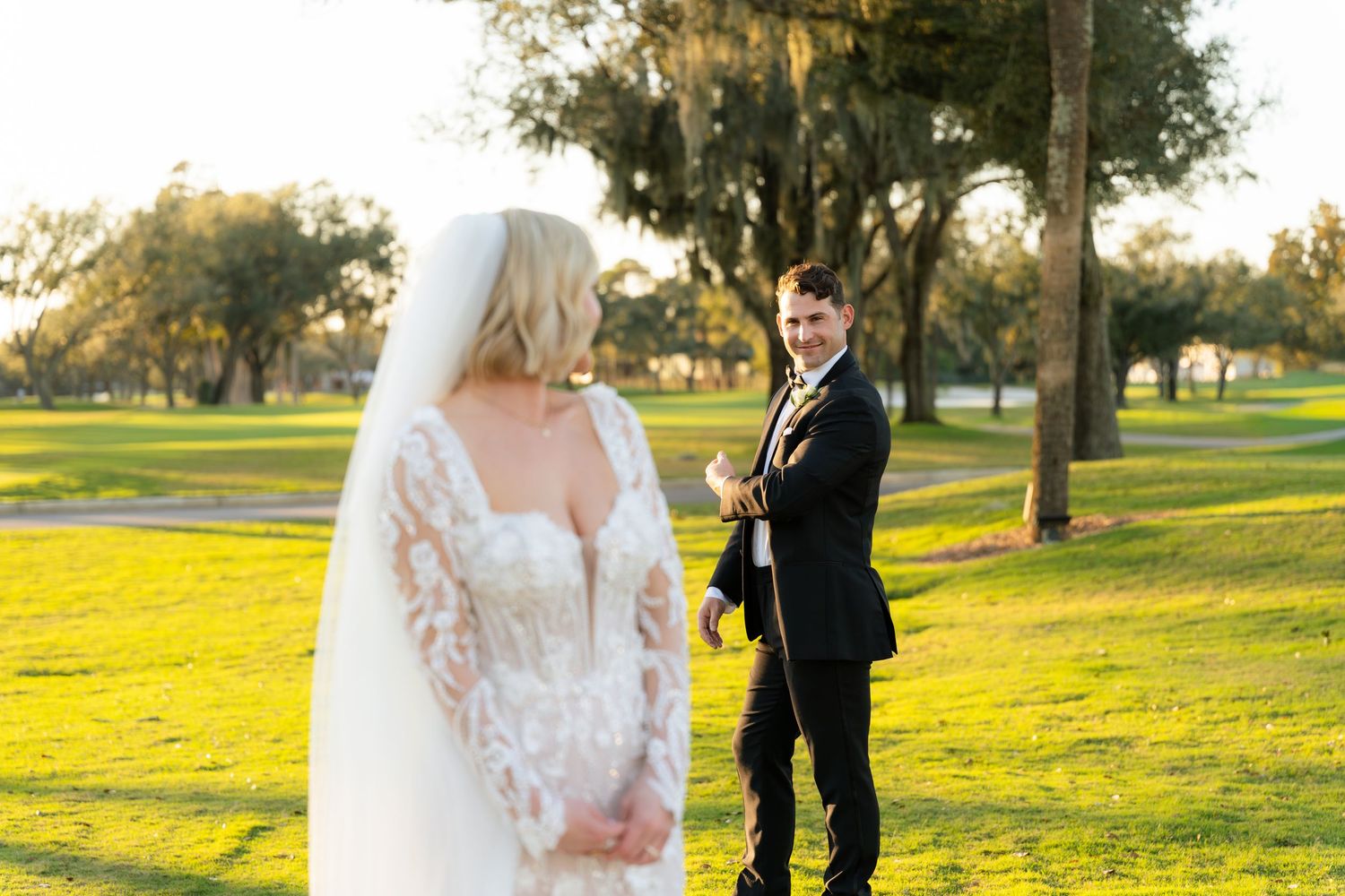 Bride in lace wedding gown with veil and groom in tuxedo posing on lush green golf course at sunset.