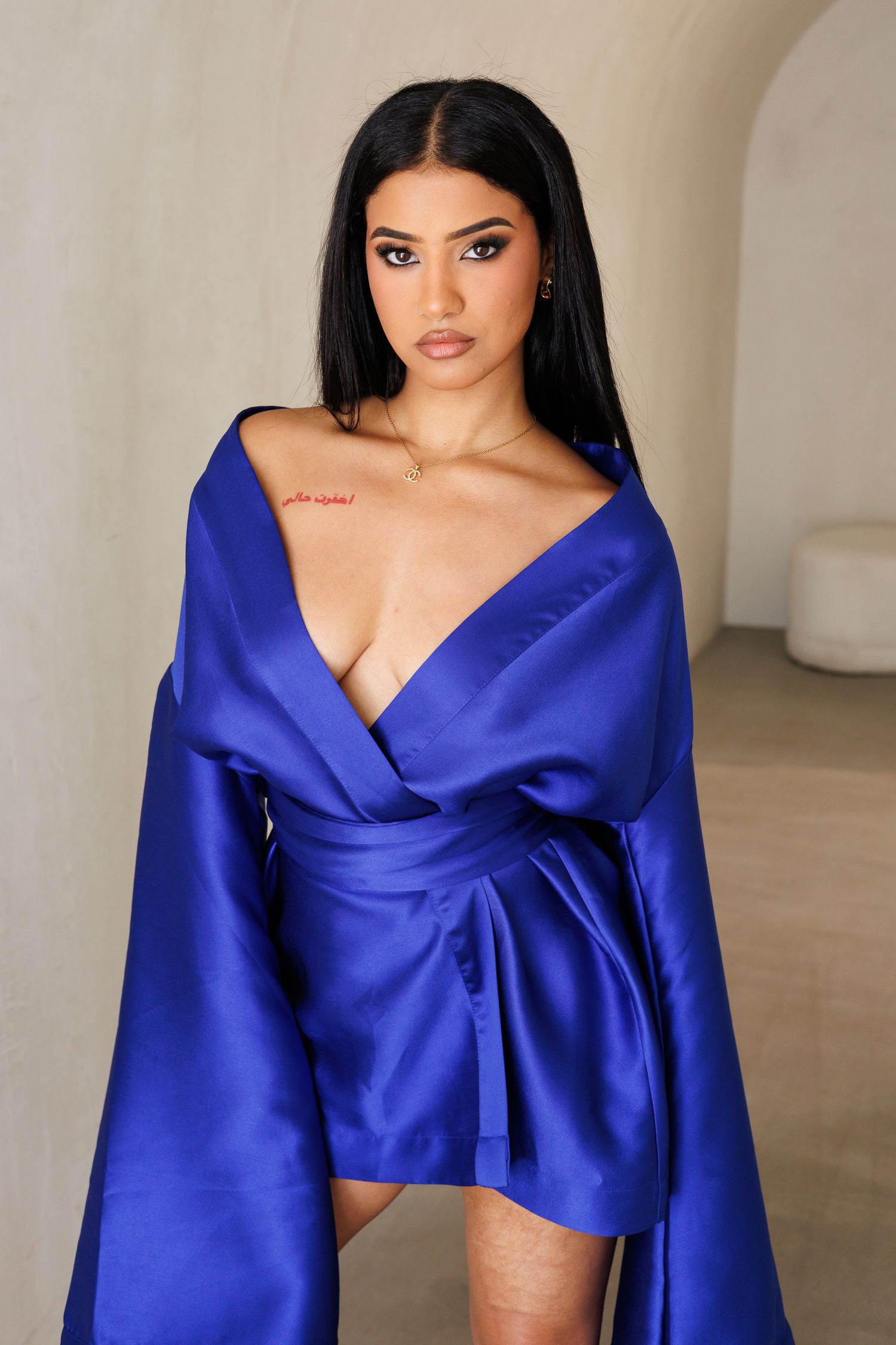 Woman in royal blue satin wrap dress with deep V-neckline and flowing sleeves, posing against neutral background.