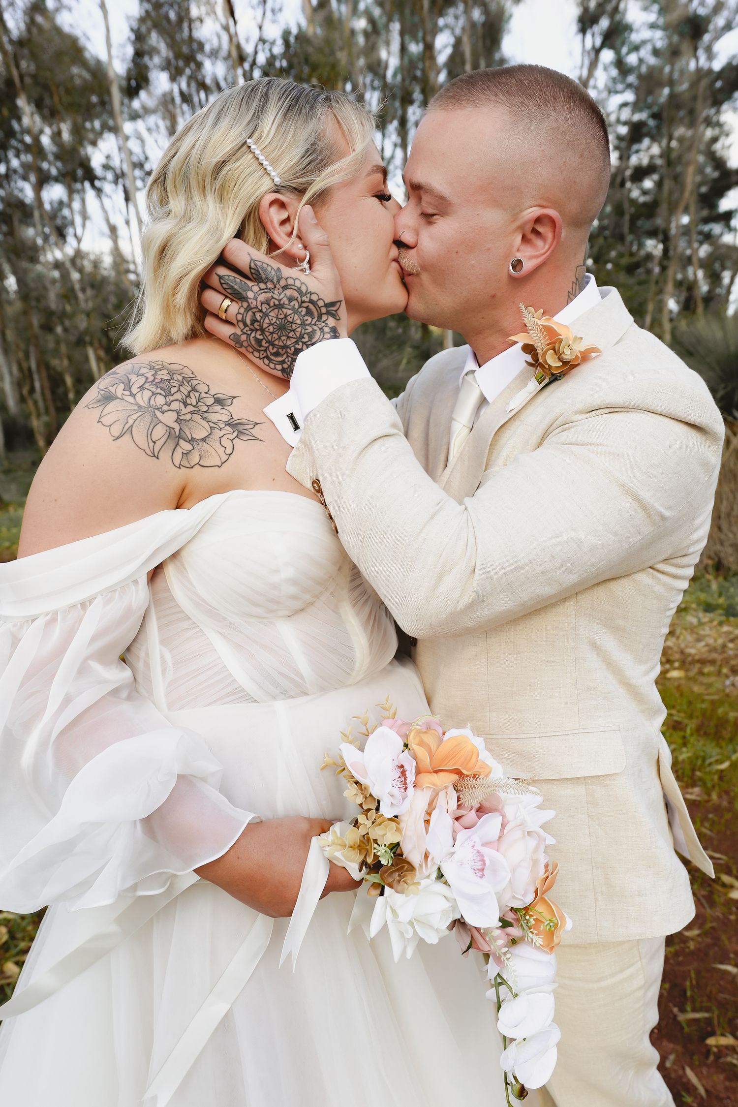Tattooed bride and groom sharing a kiss outdoors, bride holding peach and white bouquet, groom in cream suit.