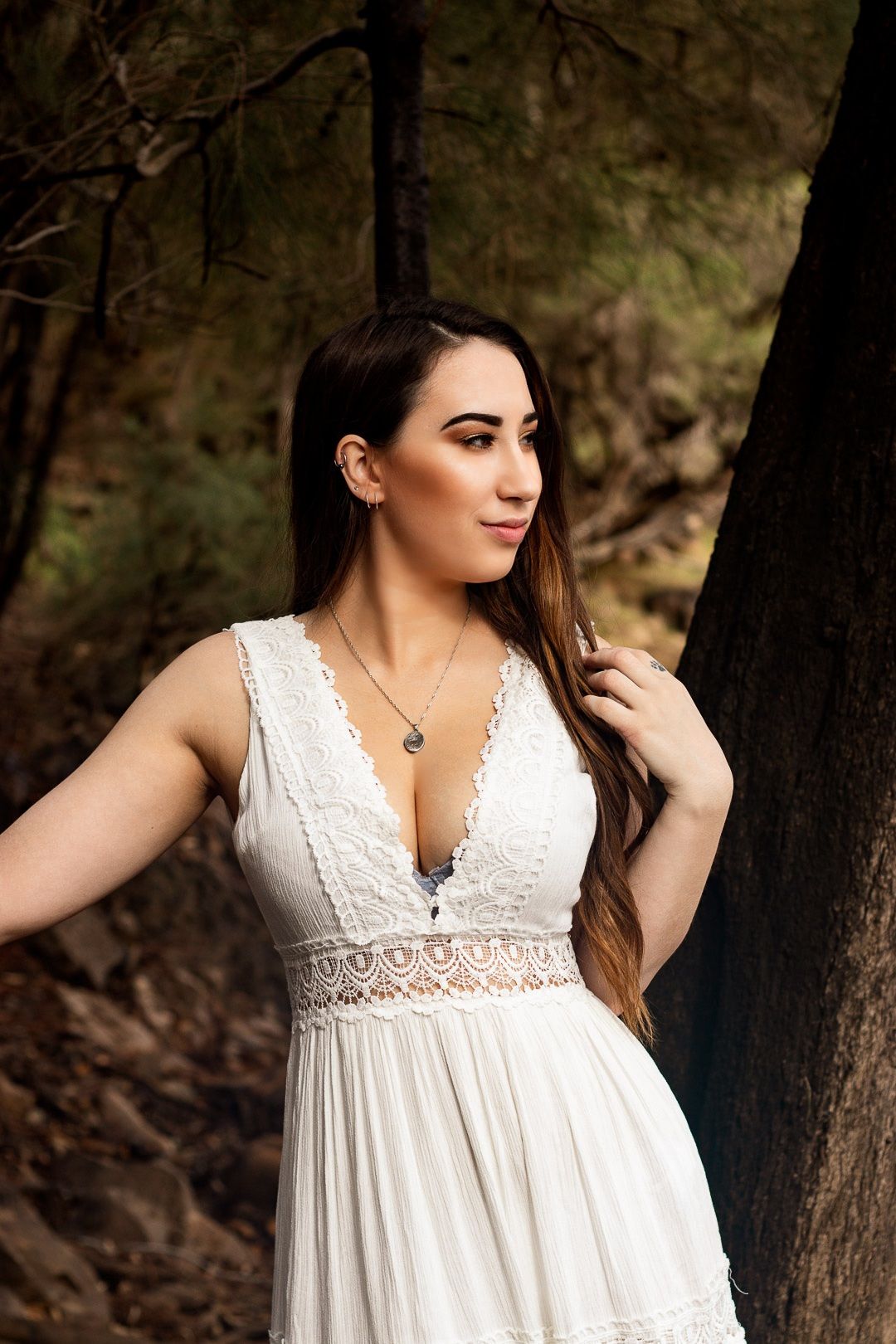 Model in white lace dress poses against forest backdrop in romantic photoshoot.