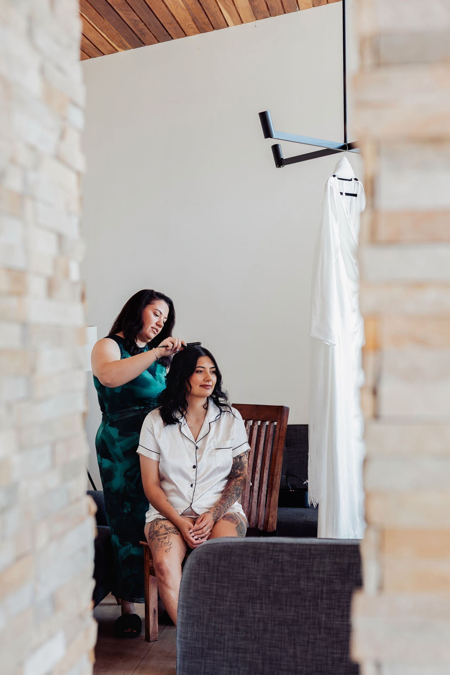 A hairstylist in a green dress styles a seated bride in pajamas, with a white wedding dress hanging in the background.