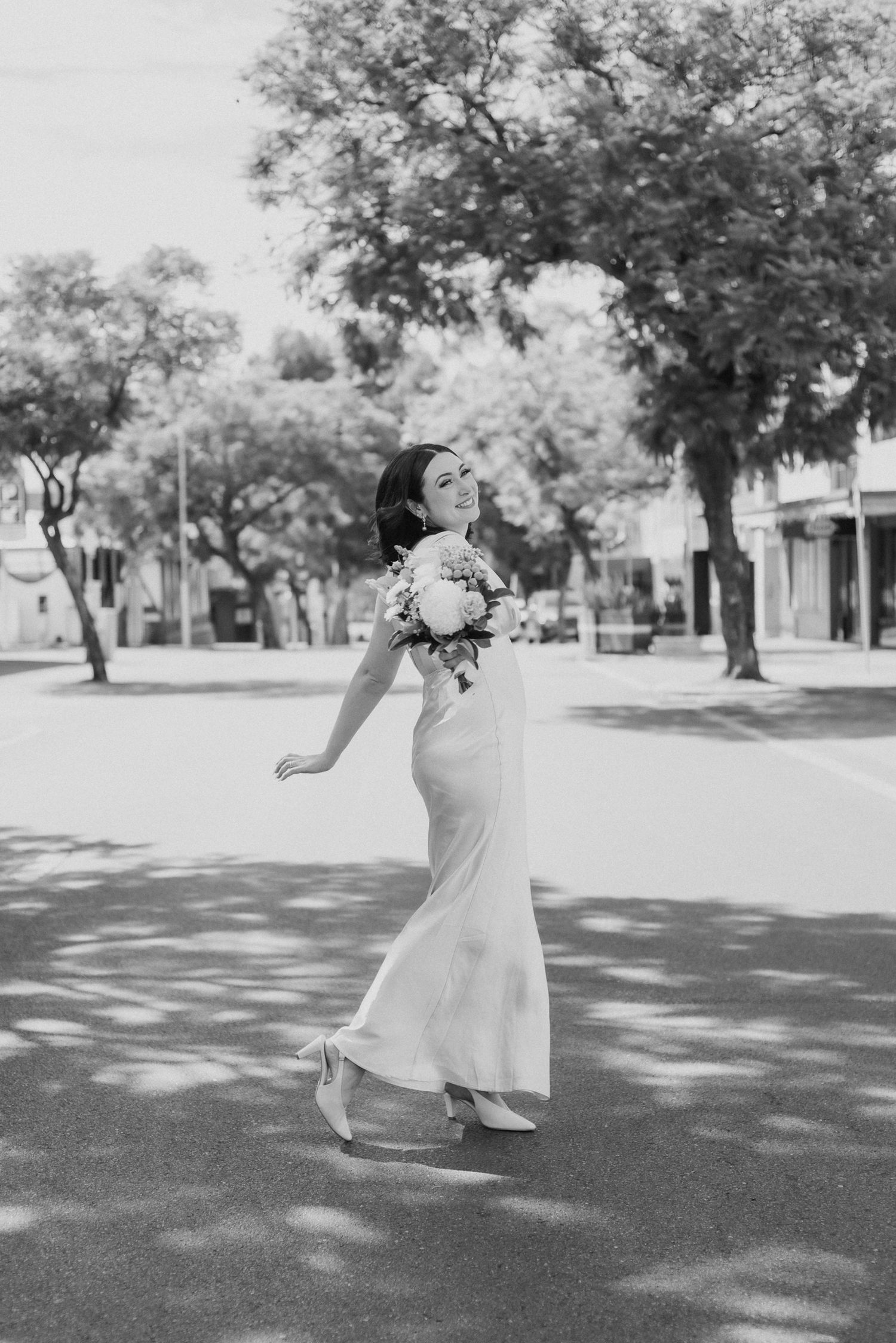 Dancing figure in white dress captured in black and white on a tree-lined path.