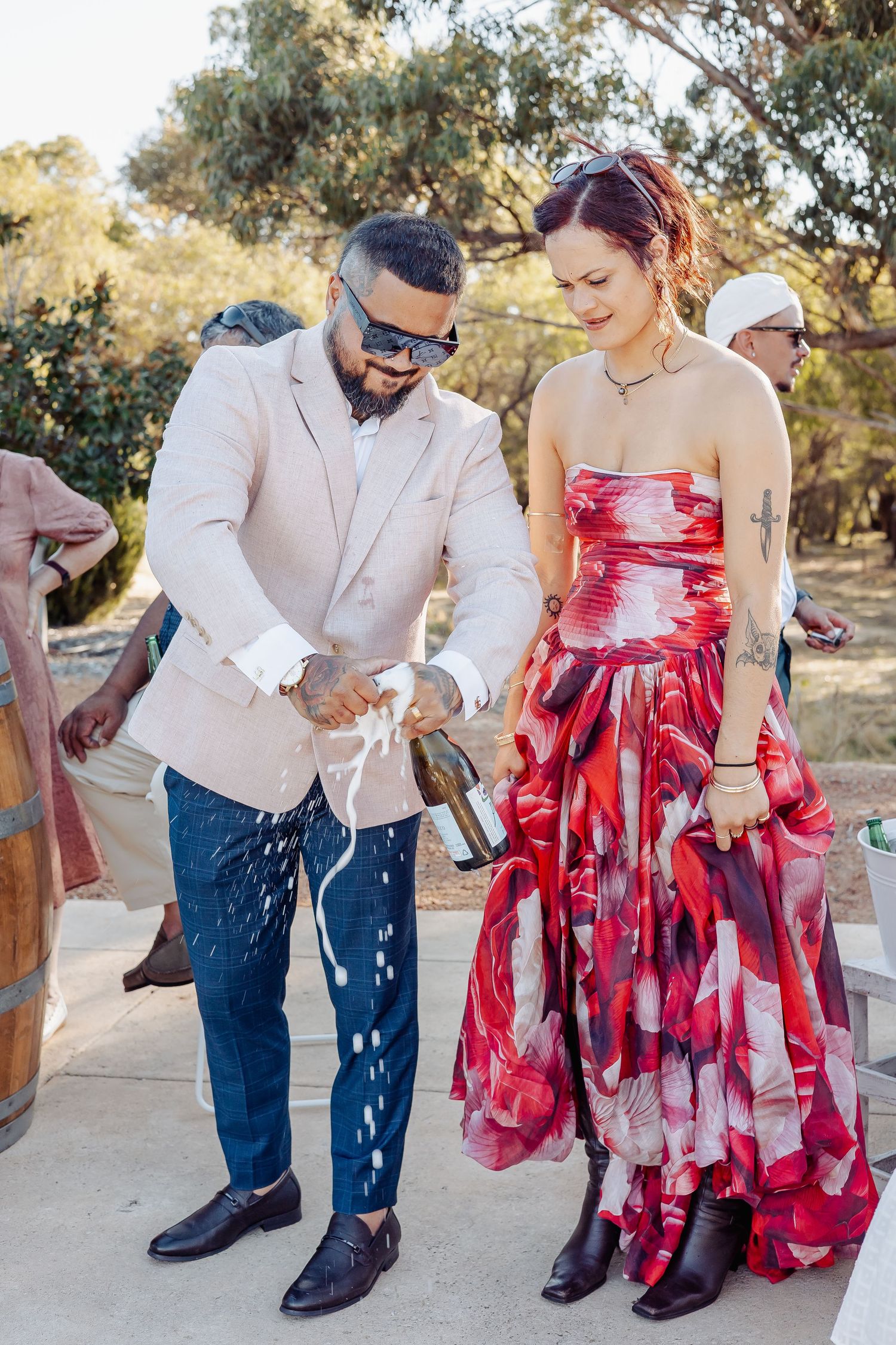 Couple in festive attire opening a champagne bottle outdoors at a celebration, man in beige suit, woman in red floral dress.
