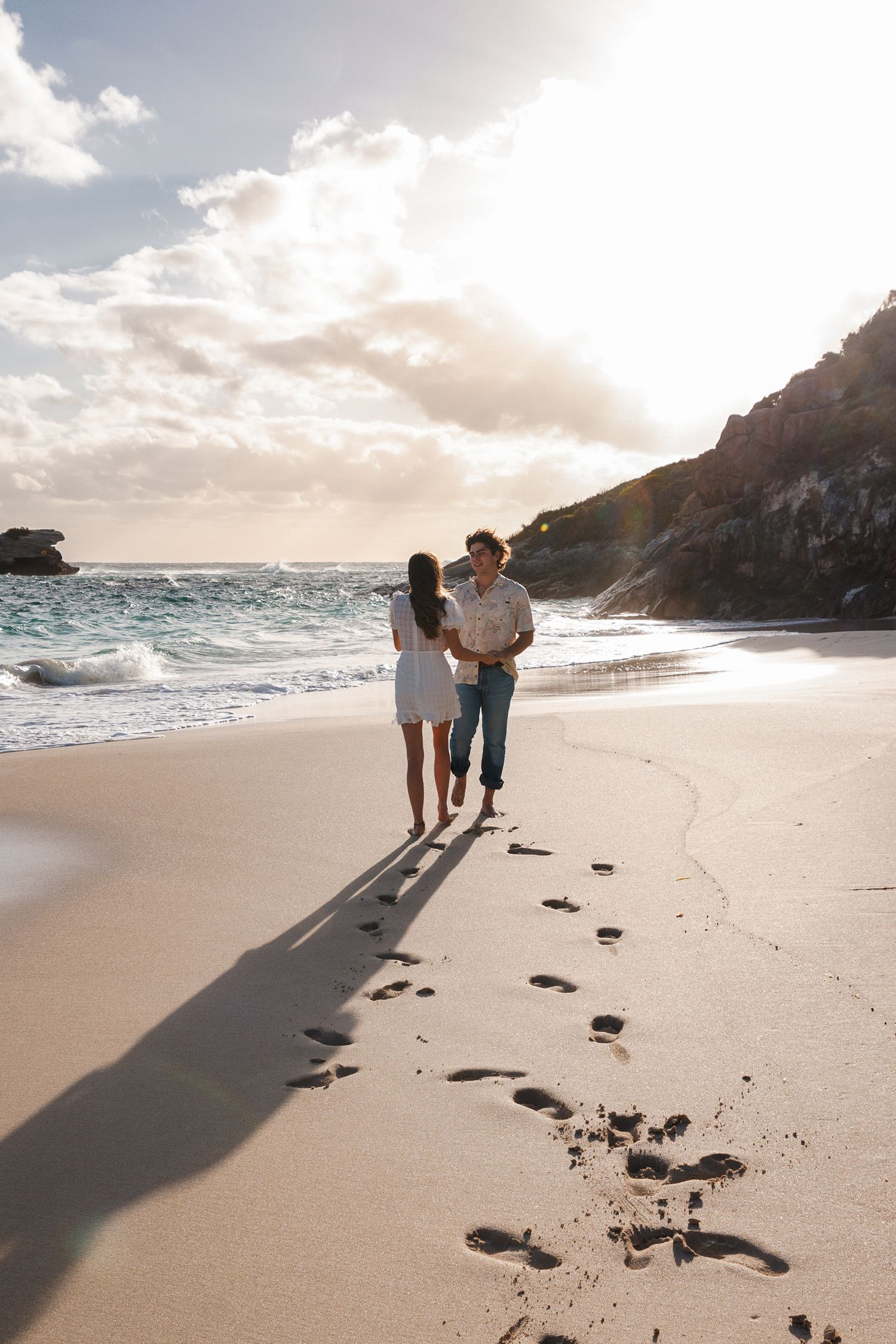 Two people walk along a scenic beach at sunset leaving footprints in the sand.