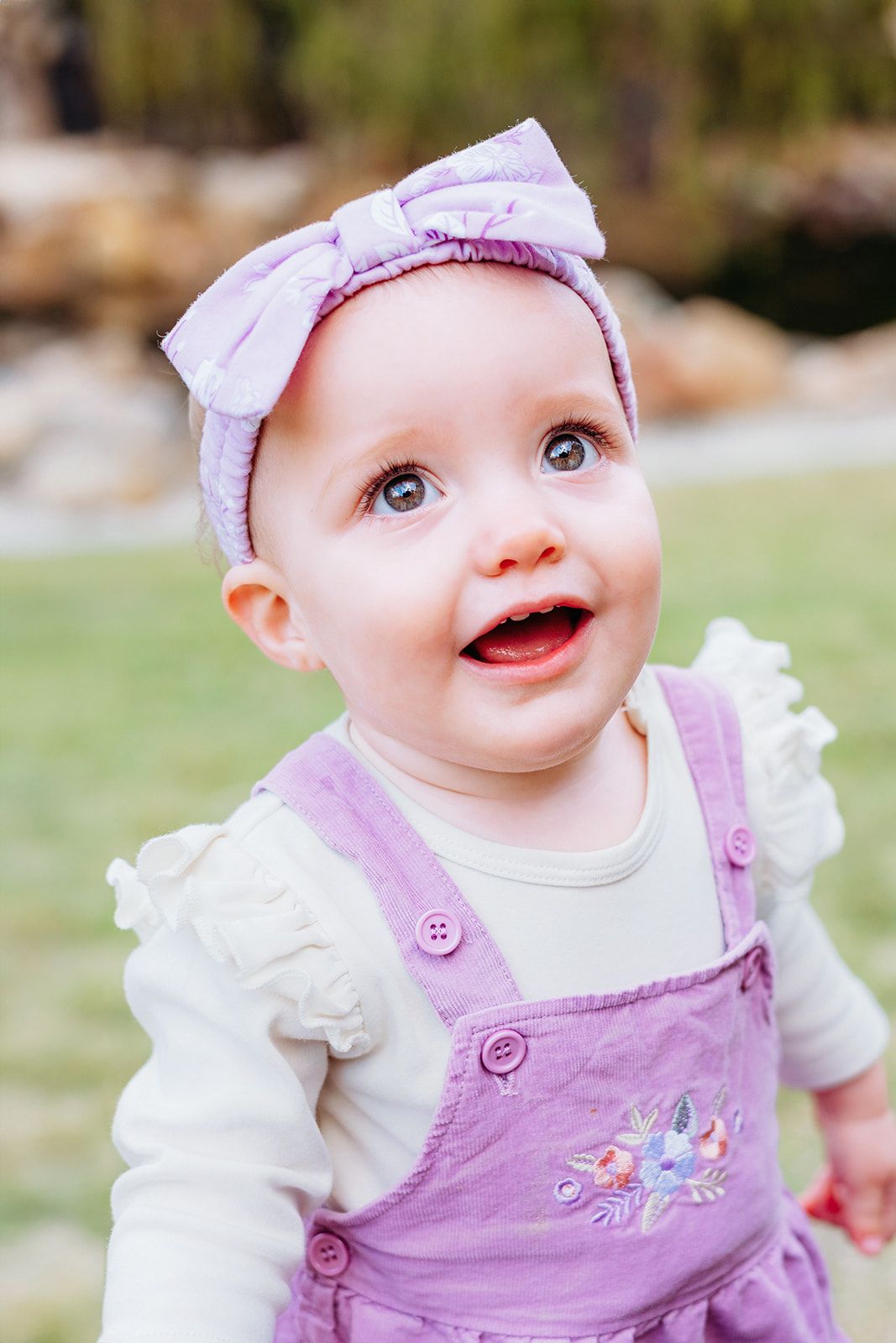 Baby in purple overalls and white bow headband smiling at camera in outdoor setting.