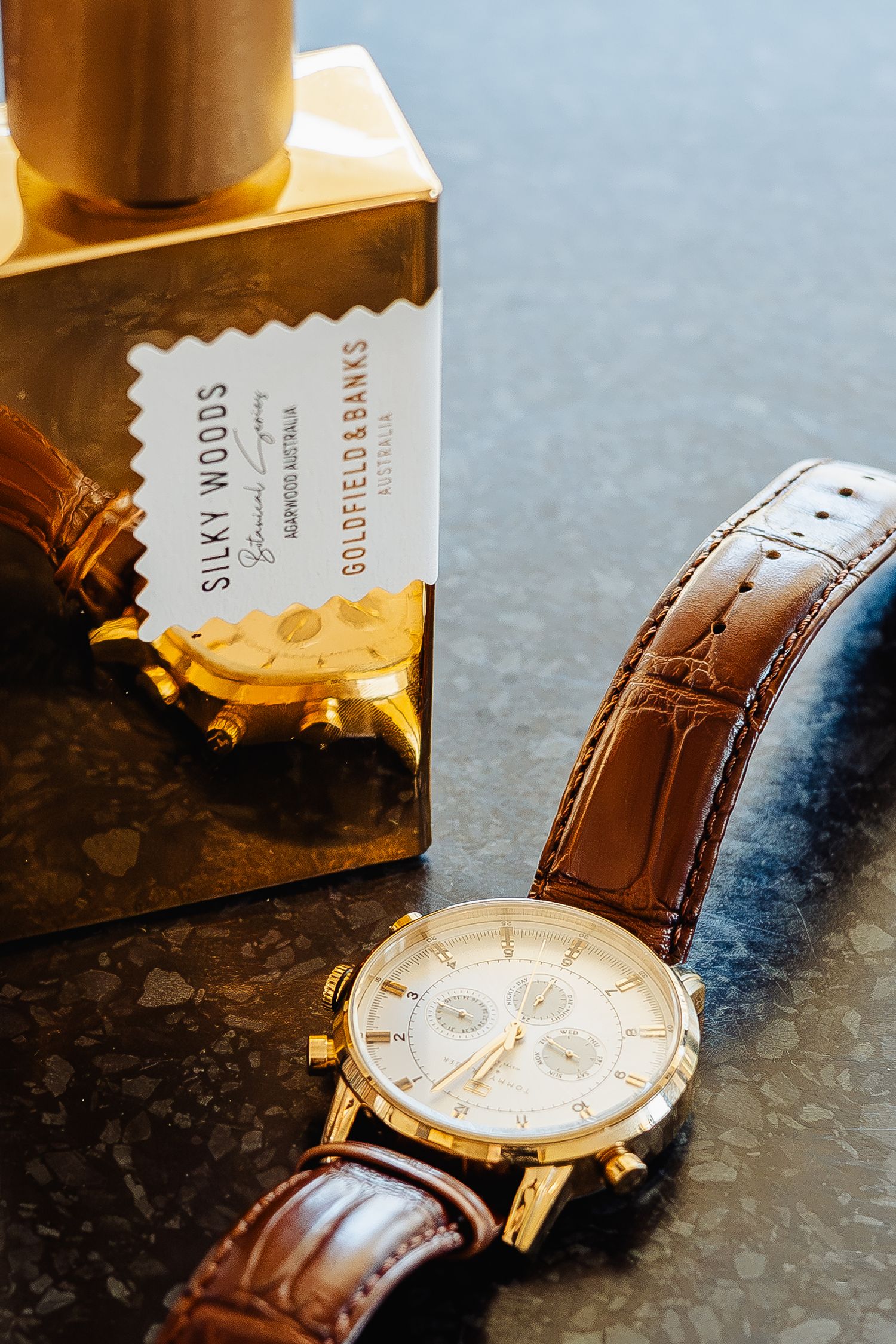 Gold chronograph watch with brown leather strap beside Goldfield & Banks Silky Woods perfume box on marble surface.