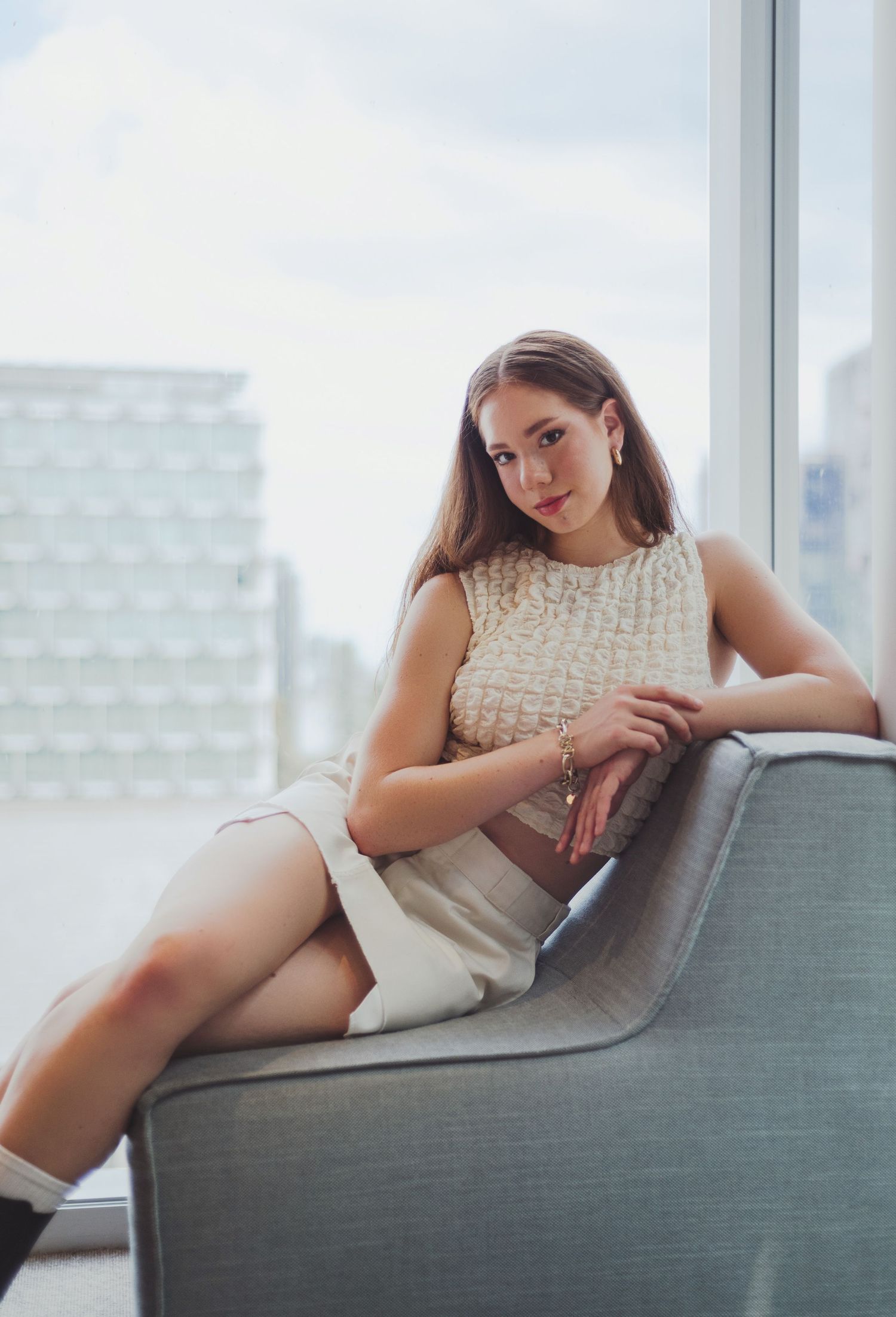 Woman in cream lace top lounges on modern gray sofa with city view behind.