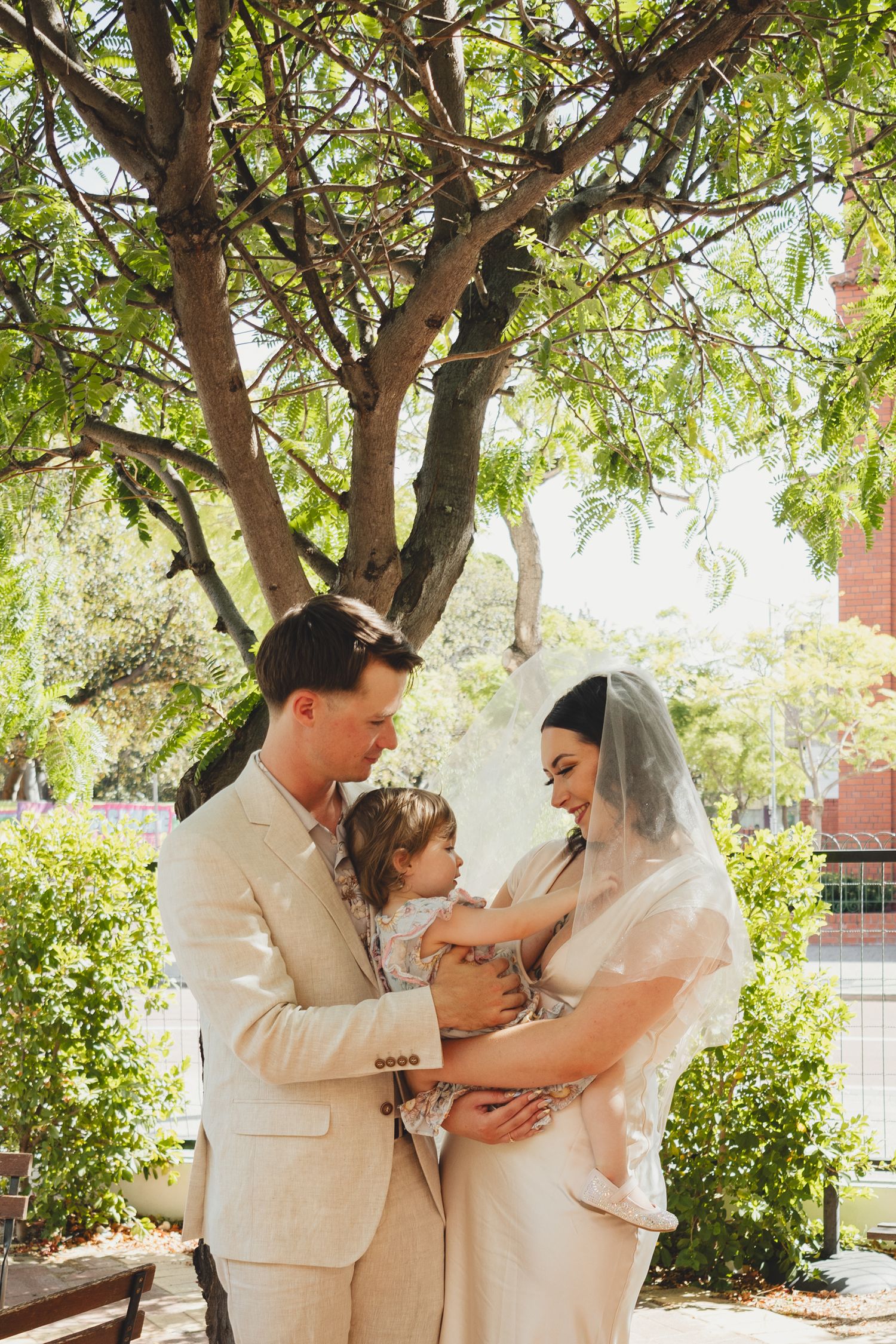 Bride and groom in neutral attire hold their young child beneath a leafy tree in a sunlit outdoor garden setting.