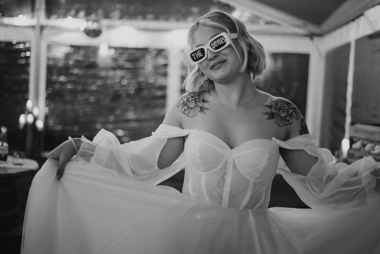 Black and white photo of tattooed bride in corset wedding gown wearing novelty sunglasses, posing elegantly outdoors.