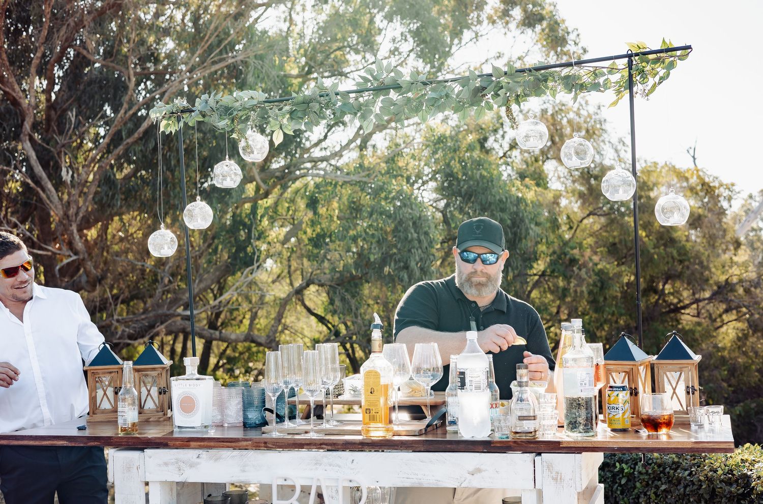 Outdoor bar setup with bartender mixing drinks under hanging globe lights and lush trees at an elegant event.