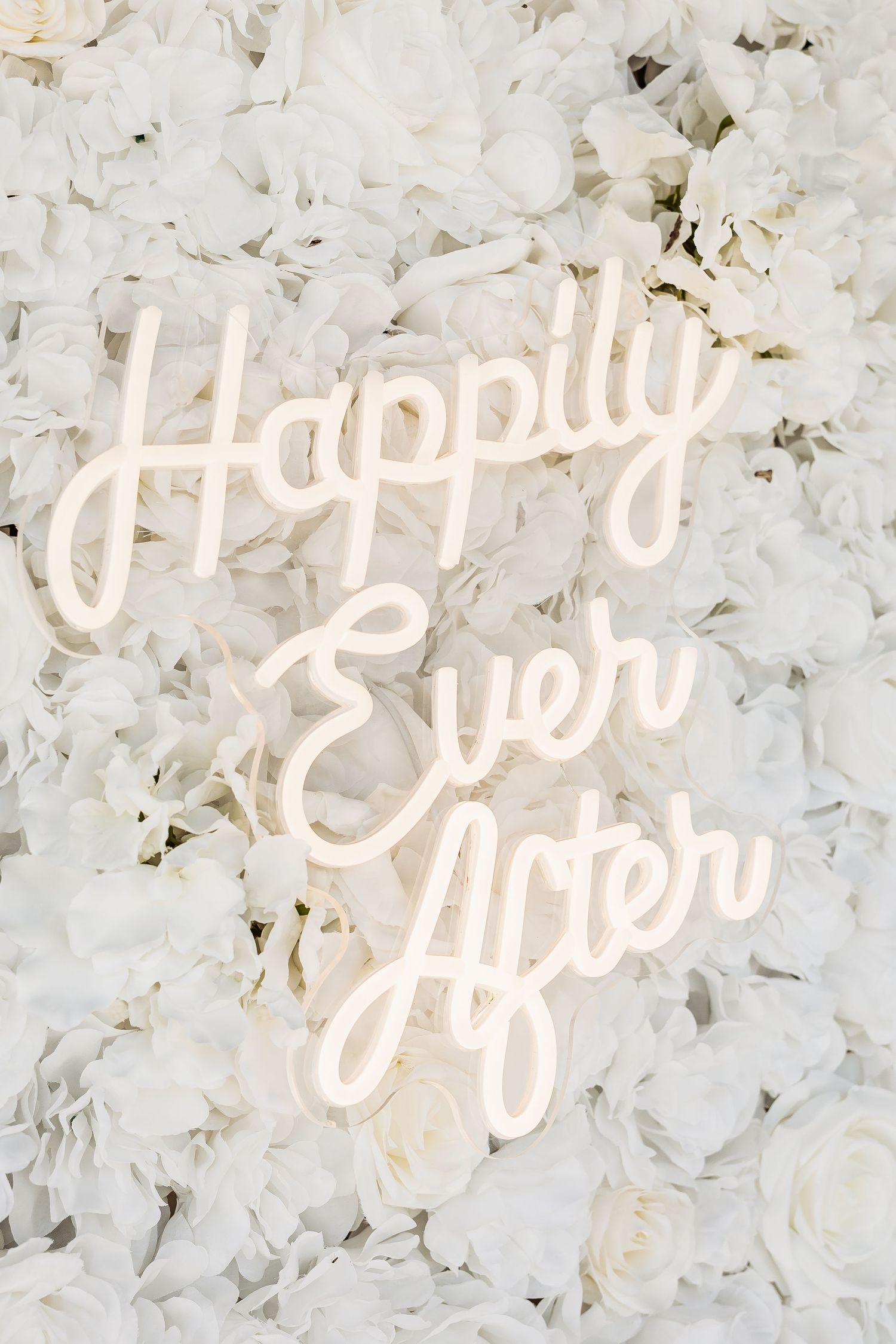 Neon sign reading Happily Ever After mounted on a white floral wall backdrop for a wedding.