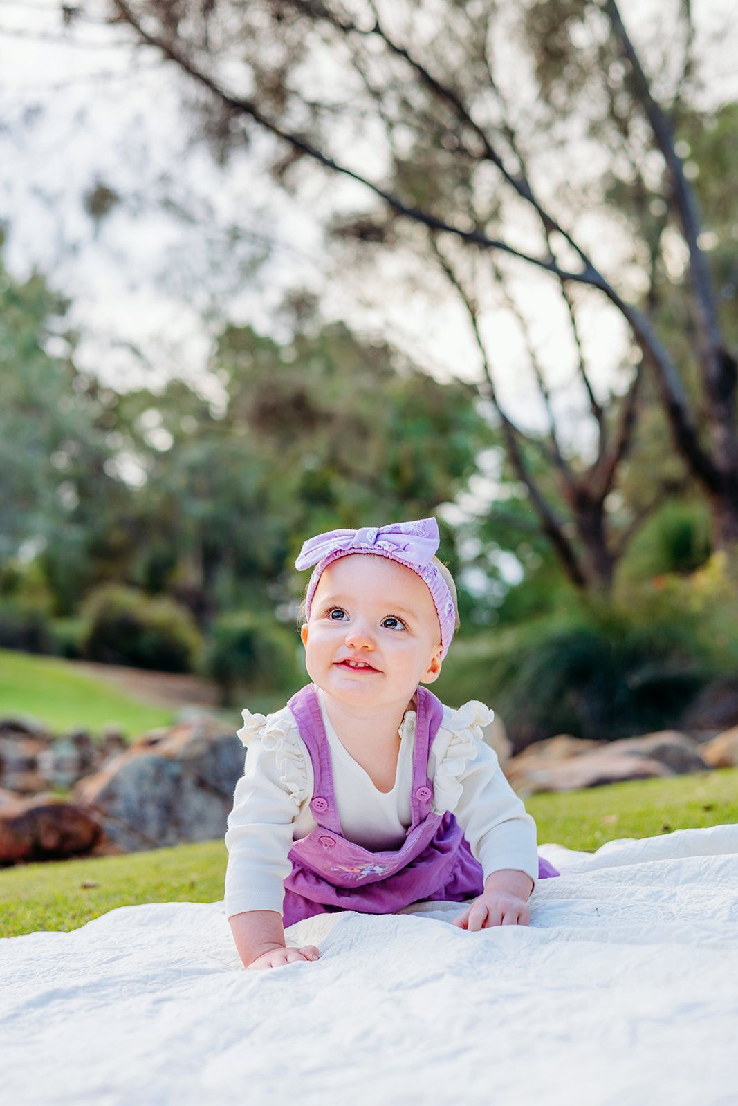 Baby in purple overalls laying on white blanket outdoors during photoshoot.